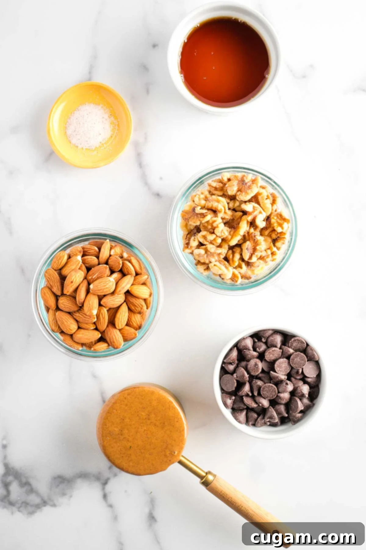 Small bowls containing the key ingredients: maple syrup, sea salt, whole almonds, whole walnuts, smooth almond butter, and dark chocolate chips.