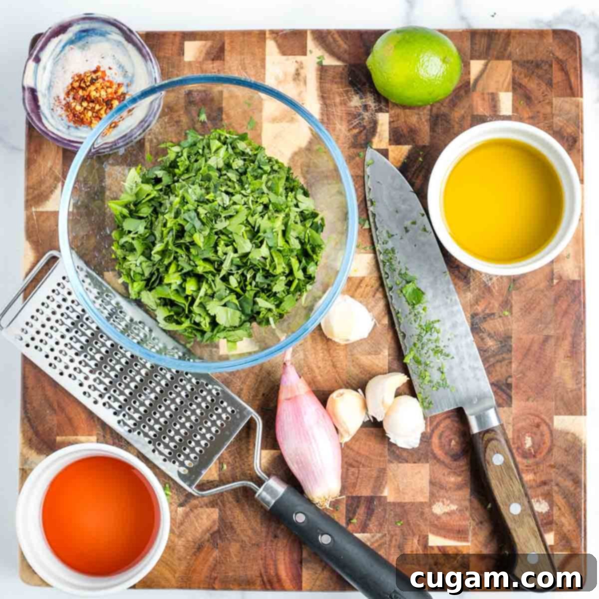 Cutting board with finely chopped fresh herbs, garlic cloves, a lime, salt and pepper, red wine vinegar, and olive oil for chimichurri.