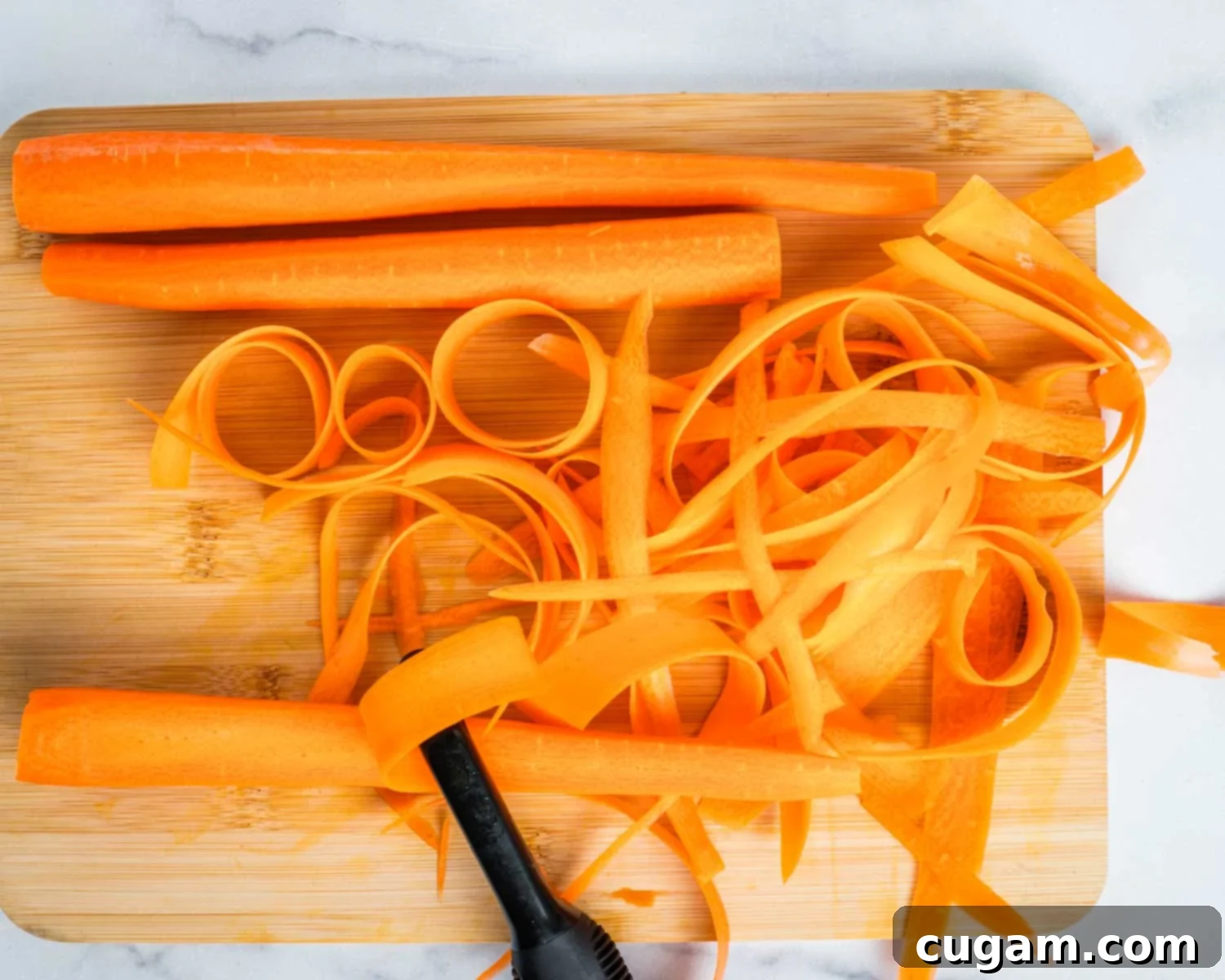 Cutting board with large carrots and a vegetable peeler pulling of a ribbon of thinly sliced carrot. A person's hand is visible holding the peeler.
