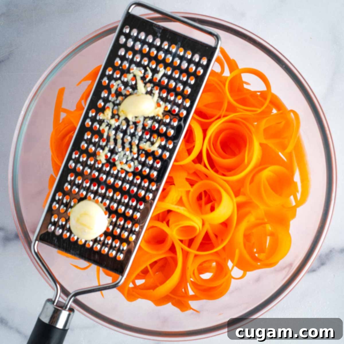 Microplane, grating fresh garlic cloves over a bowl of carrot ribbons. A chef's hand holds the microplane.