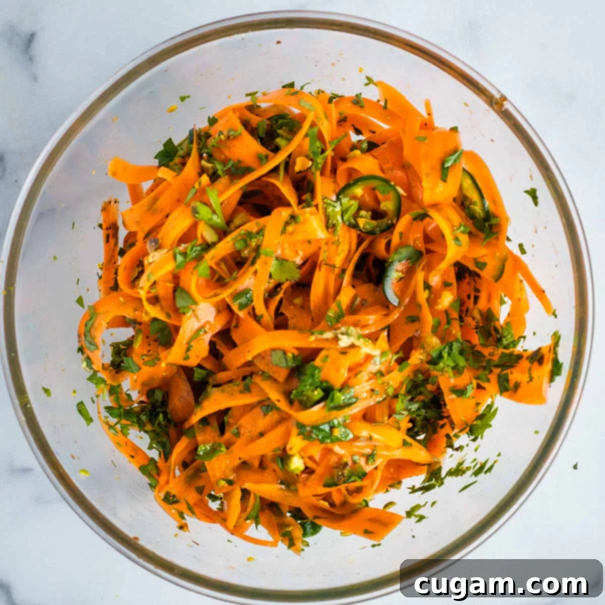 A person using tongs to toss a bowl of carrot ribbons, ensuring all ingredients are well combined. The salad appears vibrant and evenly dressed.