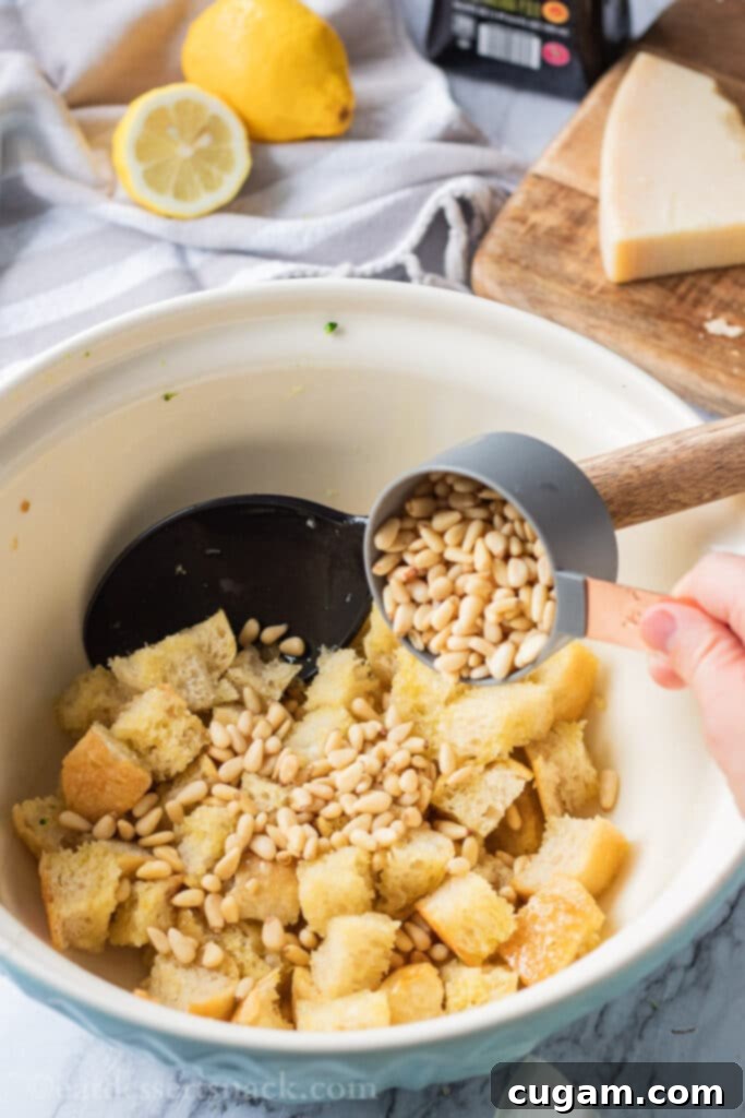 Cup pouring pine nuts into bowl of sourdough bread cubes