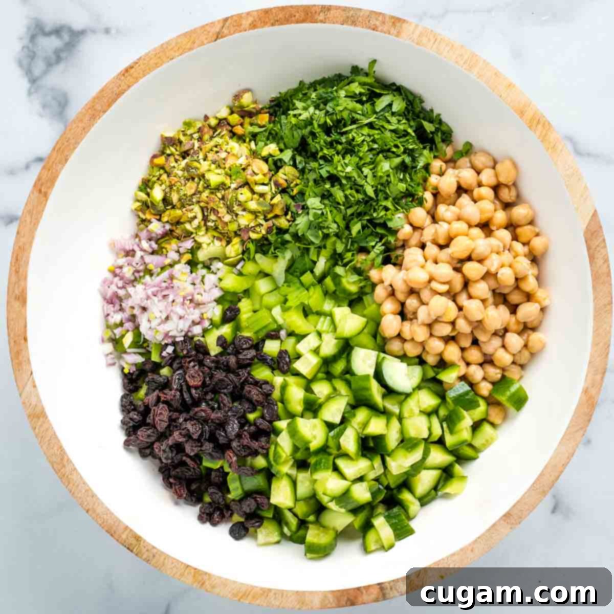 Sections of fresh herbs, chickpeas, cucumbers, raisins, pistachios and shallot in a salad bowl on top of chopped celery;.