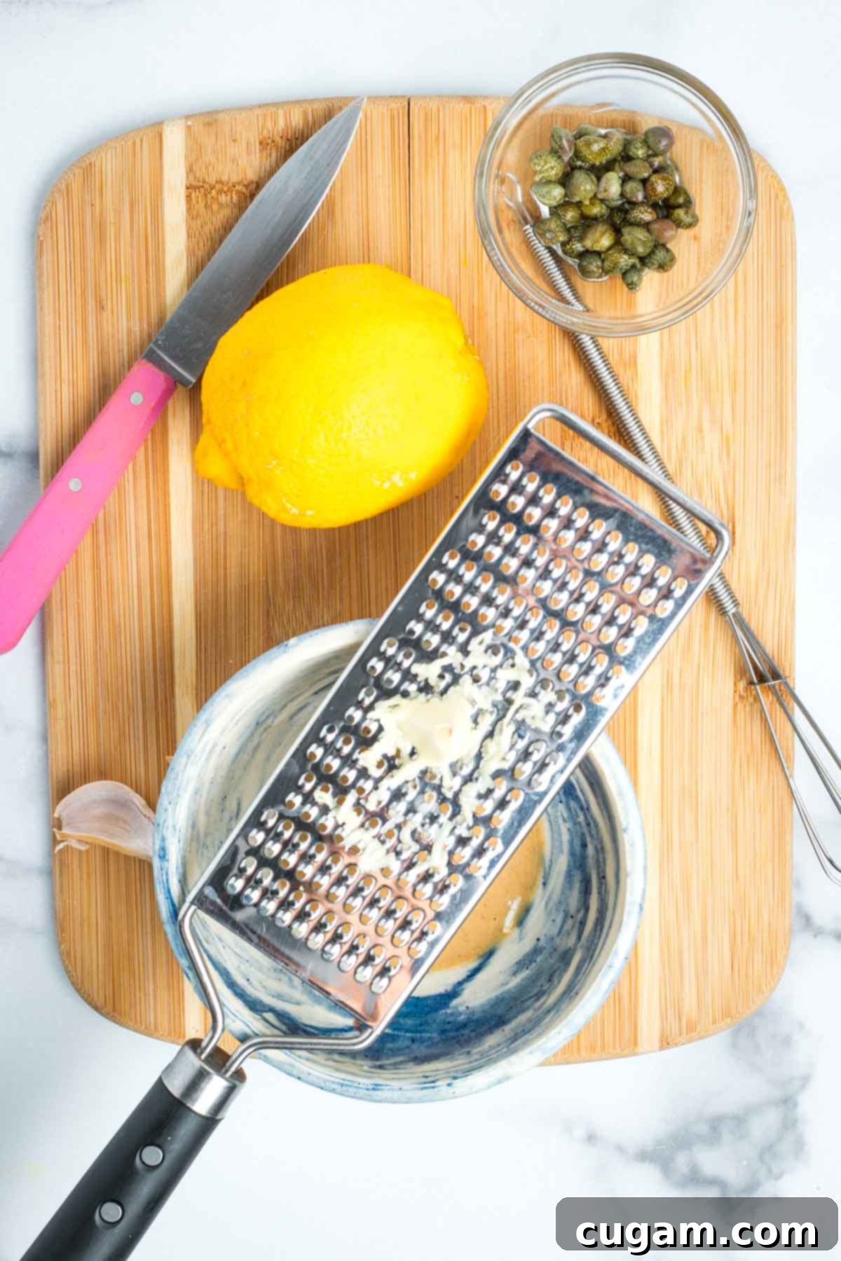 Zesting a clove of garlic over a bowl of tahini with lemon and caper on the cutting board.