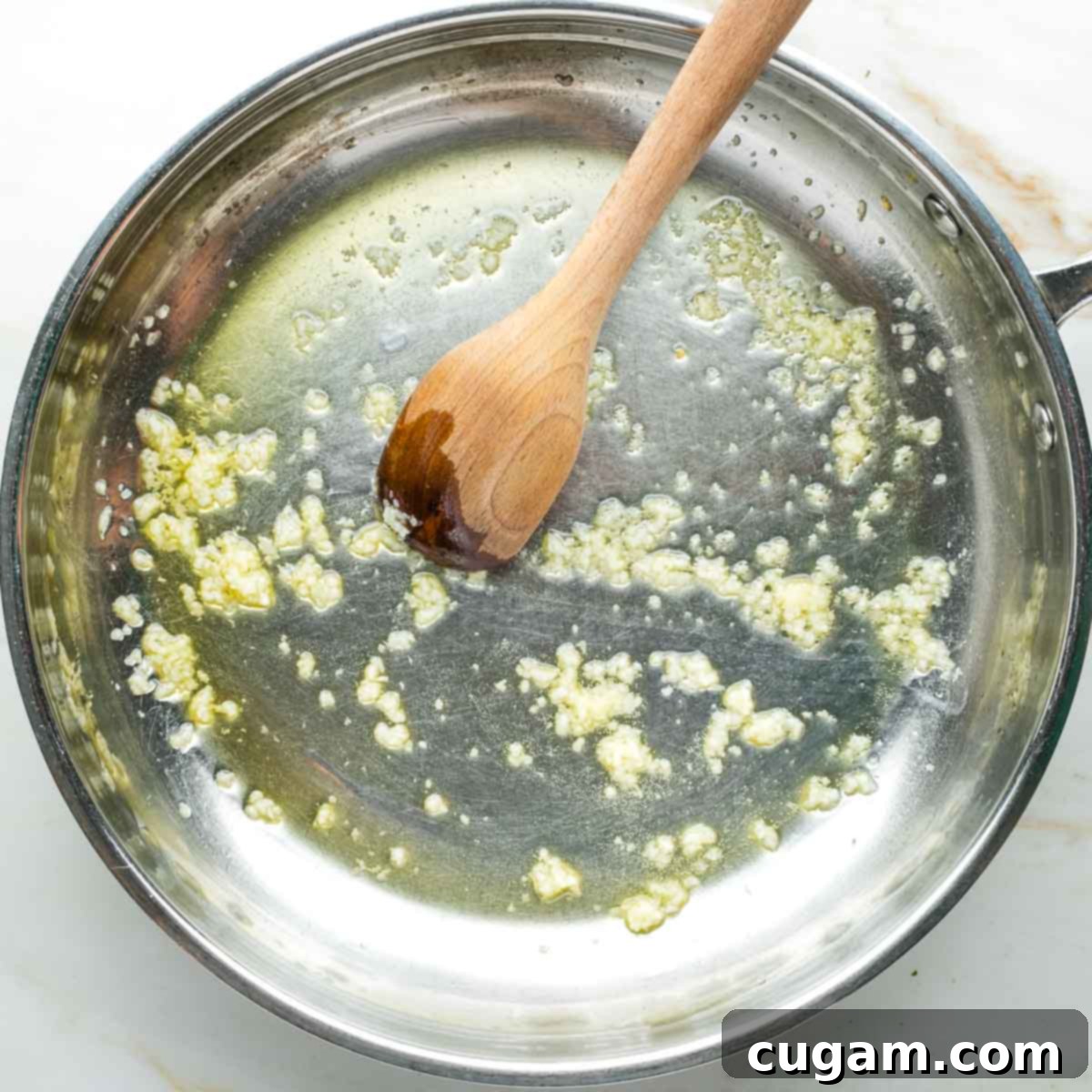 Crushed garlic gently sautéing in olive oil, being stirred by a wooden spoon in a skillet.