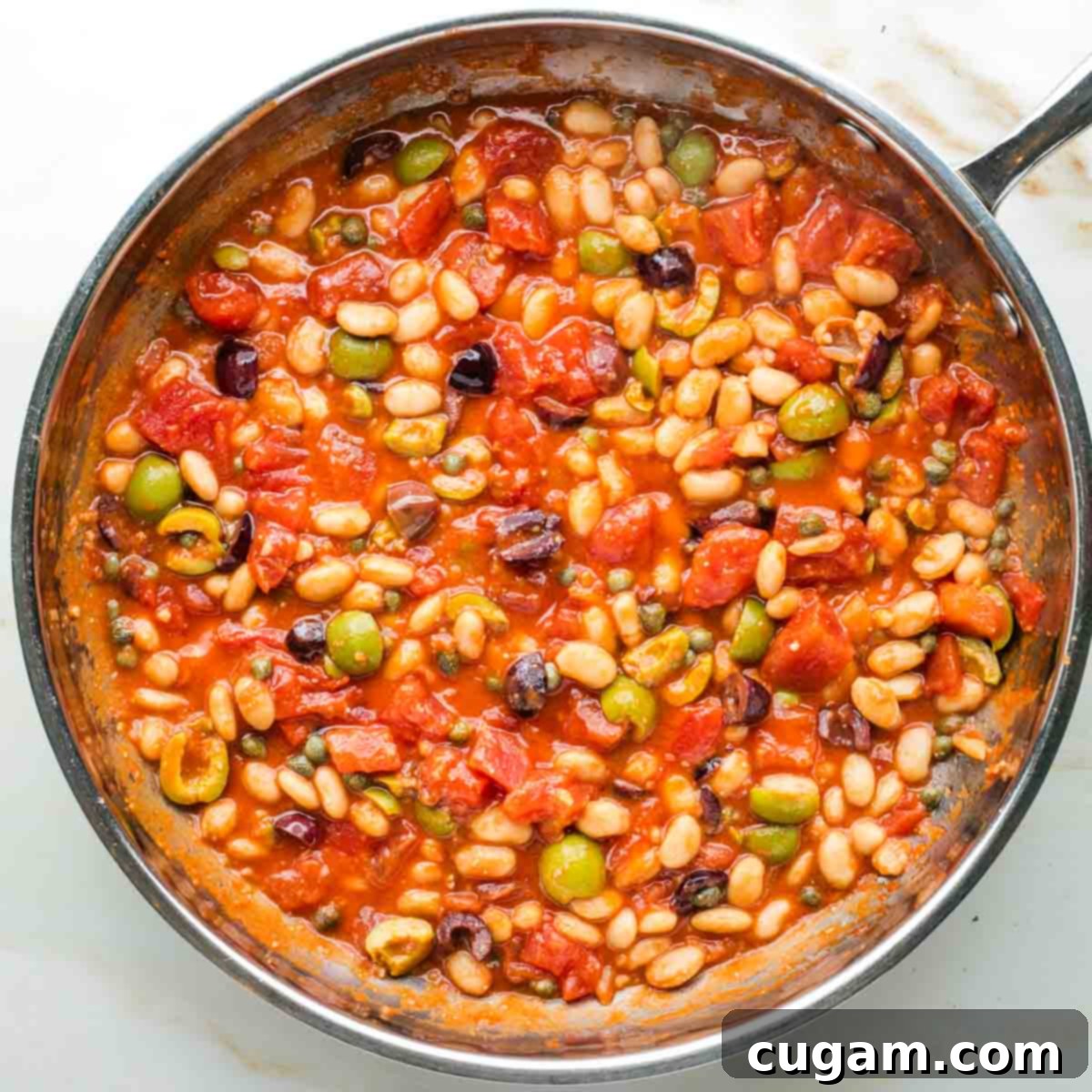 Puttanesca sauce simmering in a skillet with tomatoes, olives, capers, and white beans.