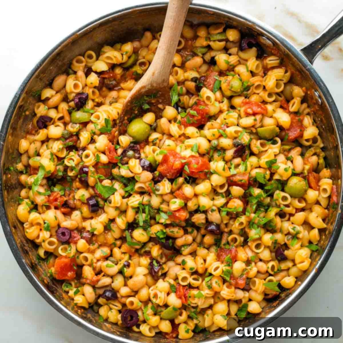 Al dente pasta being tossed with white beans, olives, capers, and fresh parsley in a chunky tomato base.