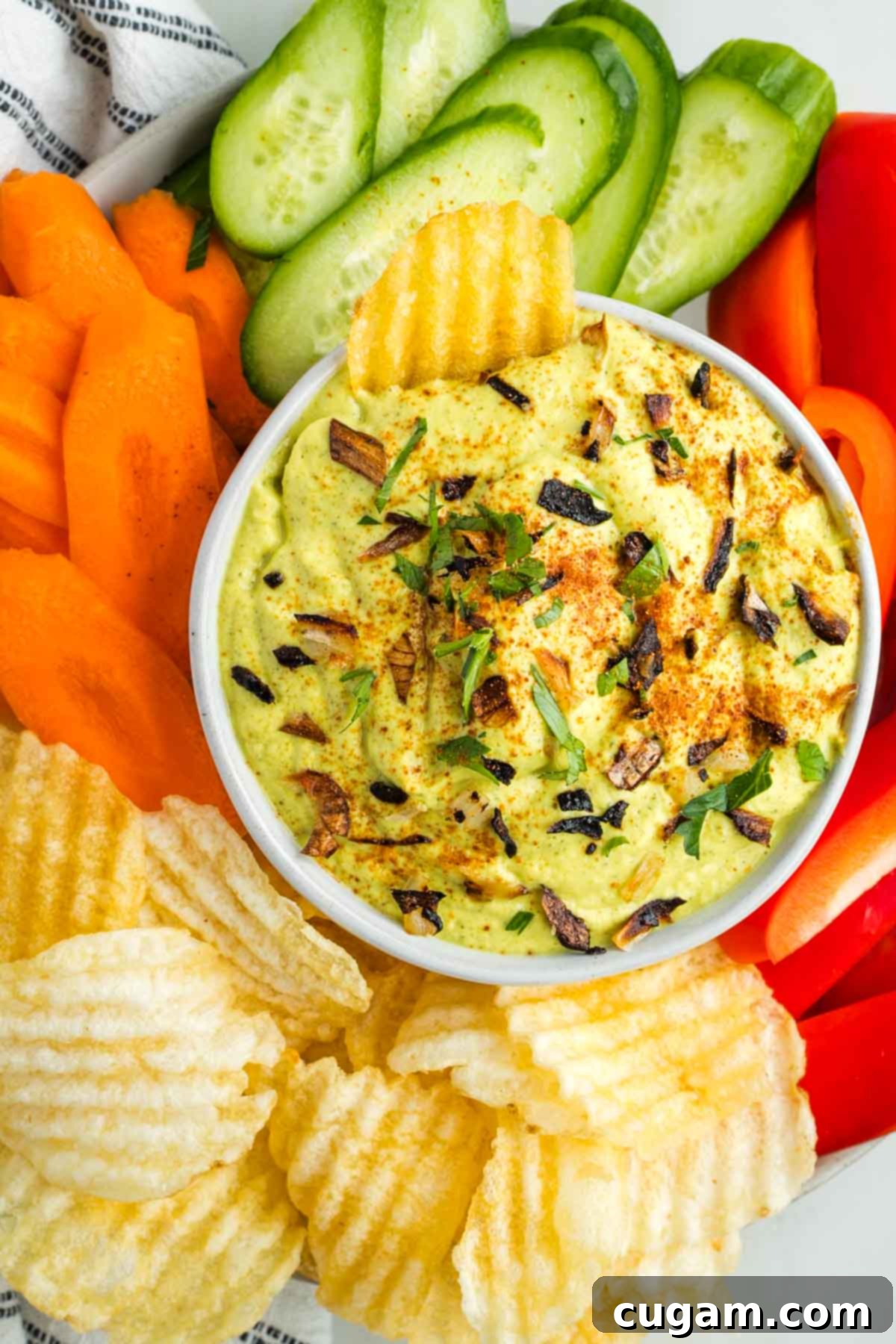 Rich, creamy vegan onion dip, speckled with golden roasted onion pieces and fresh green herbs. It's elegantly presented amidst a vibrant assortment of colorful raw vegetables like carrots, cucumbers, and bell peppers, along with a stack of crispy ridged potato chips, inviting a delightful snacking experience.