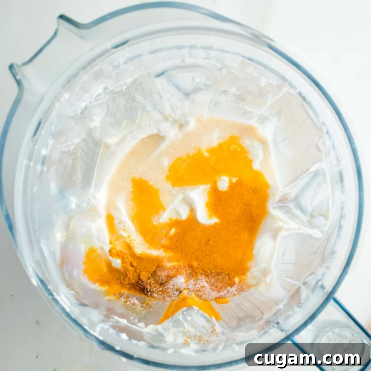 The smooth cashew cream in the blender is now sprinkled with a colorful trio of spices: vibrant turmeric, white sea salt, and a touch of fiery cayenne pepper, poised for a final blend.