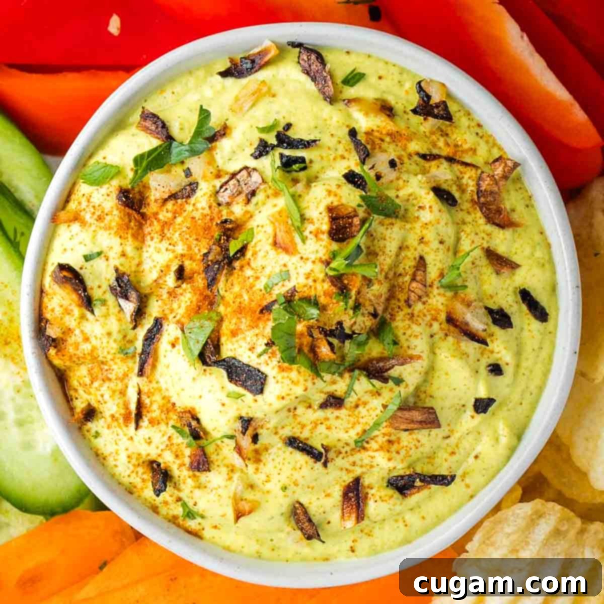 The finished creamy, light green vegan dip is elegantly presented, generously sprinkled with finely chopped crispy roasted onions, fresh vibrant parsley, and a dusting of smoked paprika for garnish. This appetizing display is ready to be served and enjoyed.