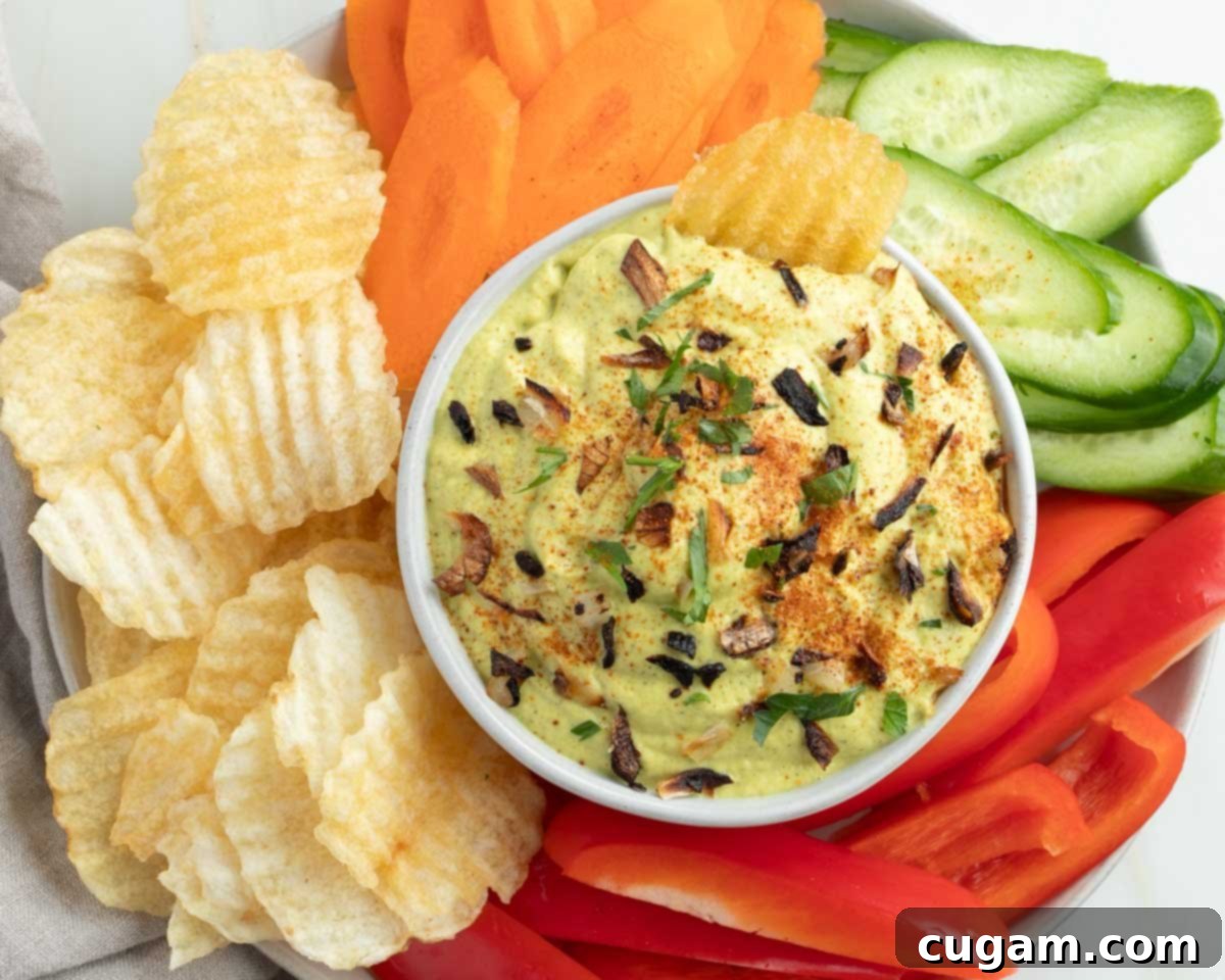 A vibrant spread featuring the vegan French onion dip, elegantly surrounded by an array of colorful fresh vegetables and a generous portion of potato chips, all arranged invitingly on a dark surface.