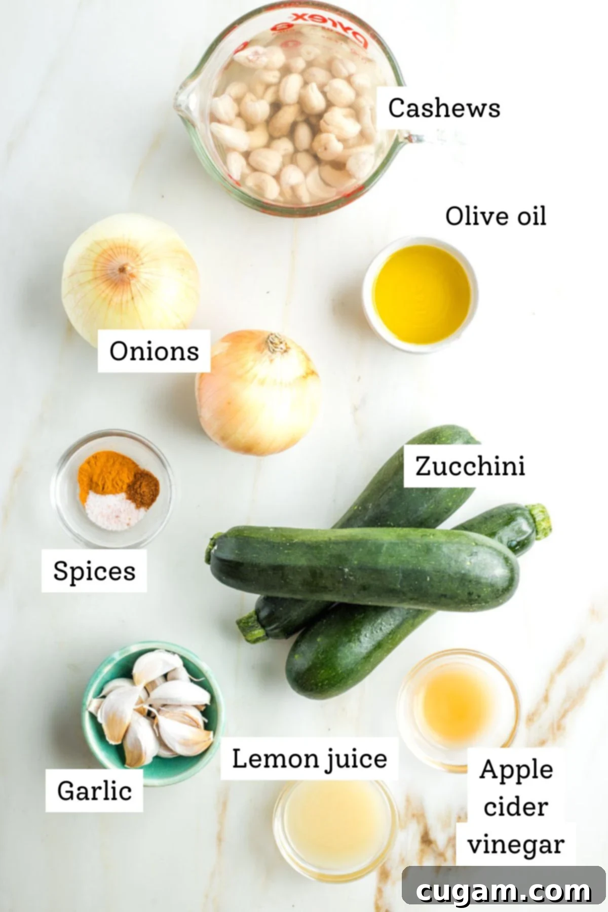 A curated selection of fresh, whole ingredients essential for the vegan roasted zucchini and onion dip, thoughtfully arranged and labeled. Featured are raw cashews, a vibrant green zucchini, a whole onion, plump garlic cloves, a bright yellow lemon, a bottle of apple cider vinegar, a small bowl of various spices (turmeric, cayenne, salt), and a cruet of olive oil, ready for preparation.