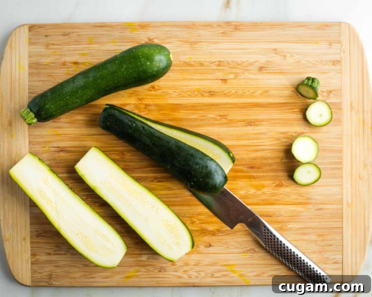 A fresh green zucchini is shown being cut in half lengthwise on a wooden cutting board, revealing its interior texture and preparing it for roasting.