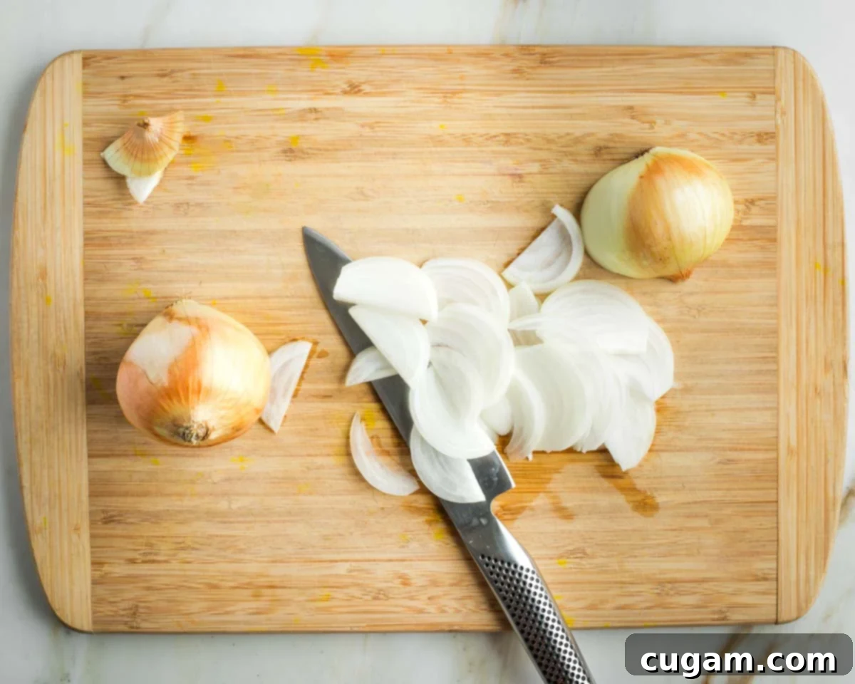 A skilled hand demonstrates how to thinly slice an onion on a wooden cutting board, ensuring even pieces for optimal roasting.