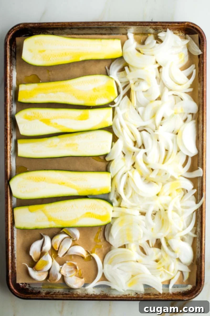 Long halves of zucchini, thinly sliced onions, and whole garlic cloves are artfully arranged on a parchment-lined rimmed baking tray, lightly drizzled with olive oil, ready for roasting.