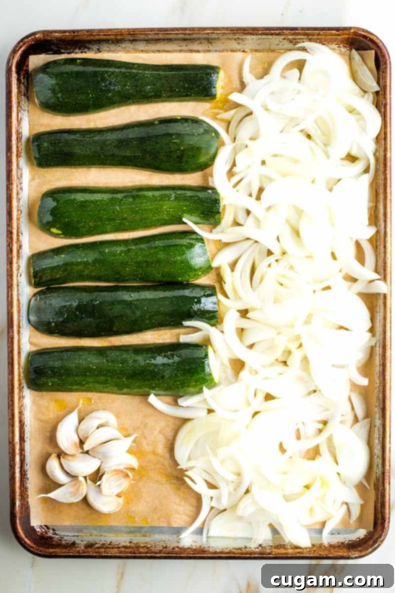 The prepared zucchini, sliced onions, and whole garlic cloves are thoroughly coated with olive oil on a parchment-lined baking tray, arranged for even roasting.