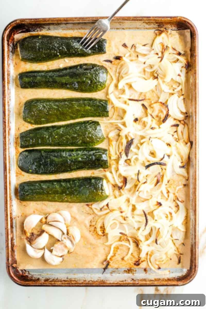 Perfectly roasted halved zucchini, tender sliced onions, and soft garlic cloves on a baking tray lined with parchment paper, indicating they are ready for the next step in dip preparation.