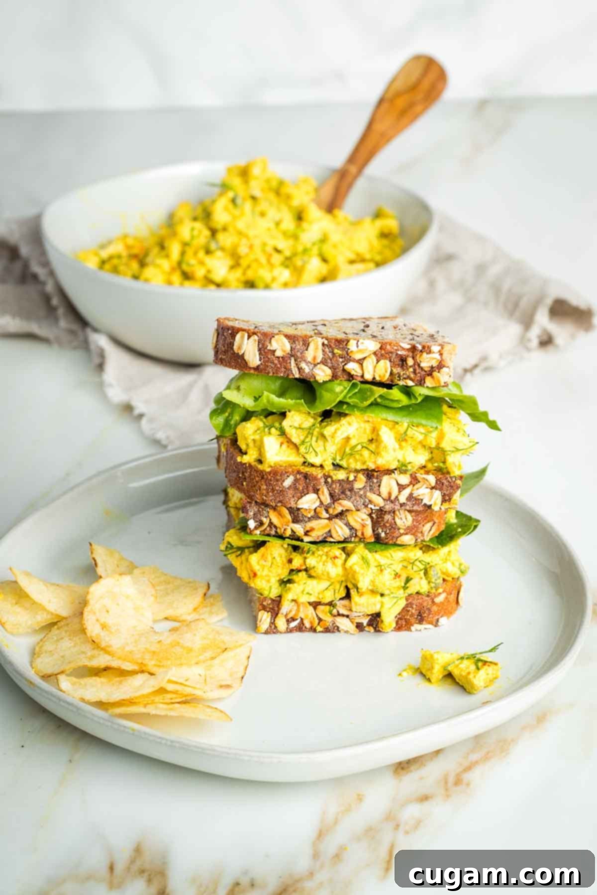 Stacked vegan egg salad sandwich with chips on the plate and a bowl of the egg salad beyond.