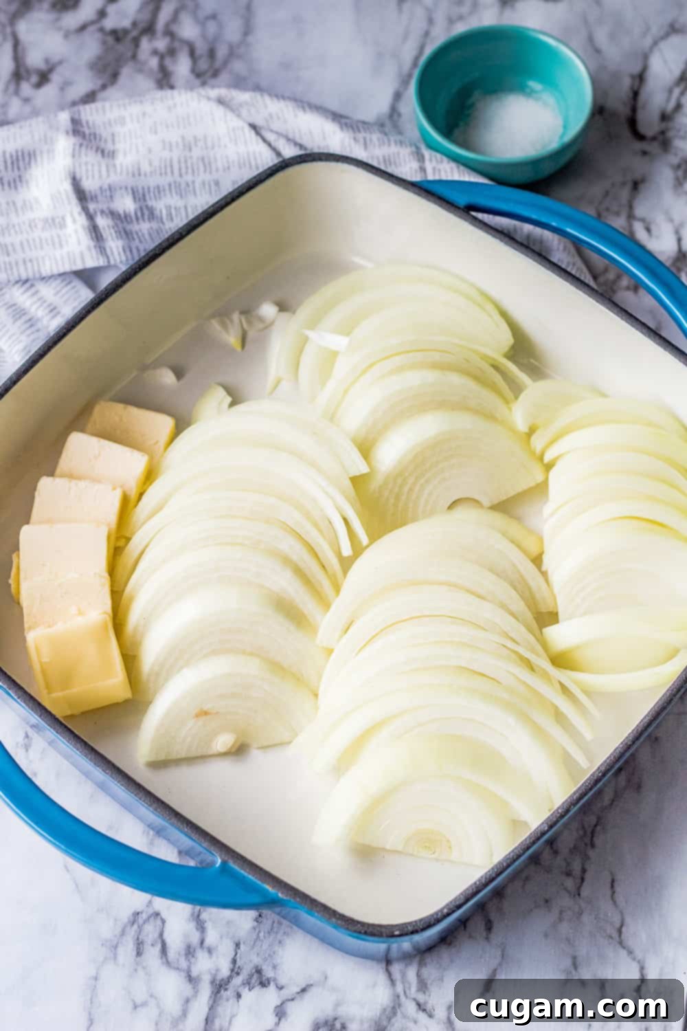 Sliced raw onions with butter in blue square pan on marble. 