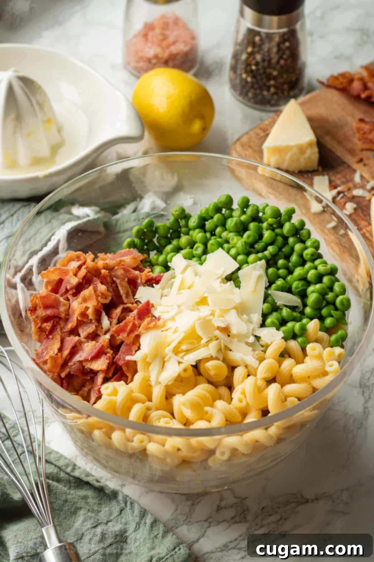 Clear glass bowl filled with cooked pasta, green peas, crispy bacon pieces, and fresh Parmesan flakes on a marble countertop.
