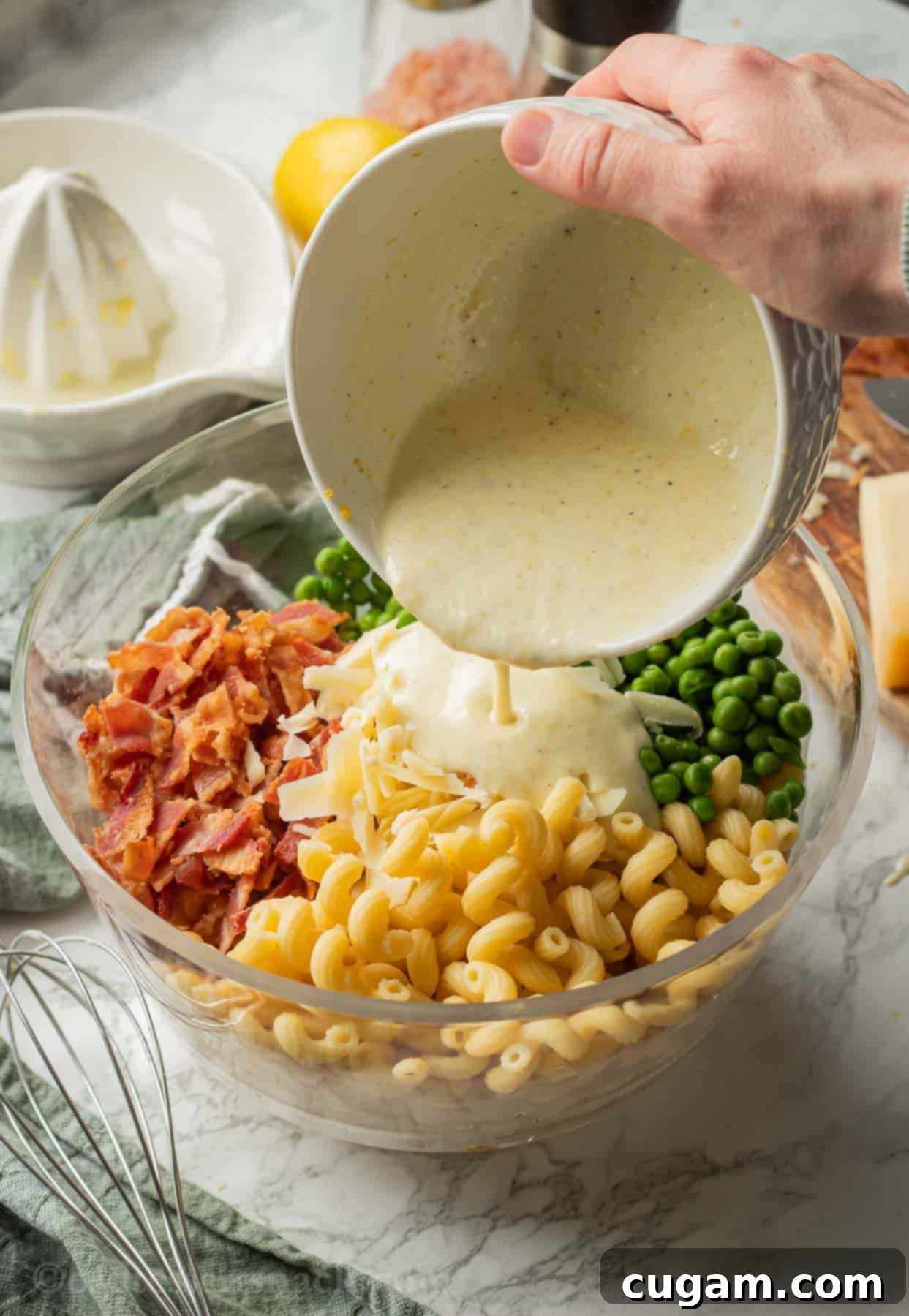 Close-up of creamy lemon dressing being poured from a small bowl over the assembled lemon pasta salad ingredients in a large bowl.