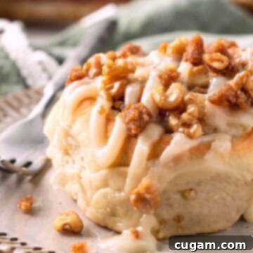 A single, perfectly baked banana cinnamon roll, generously topped with cream cheese icing, served on a white plate with a fork beside it.