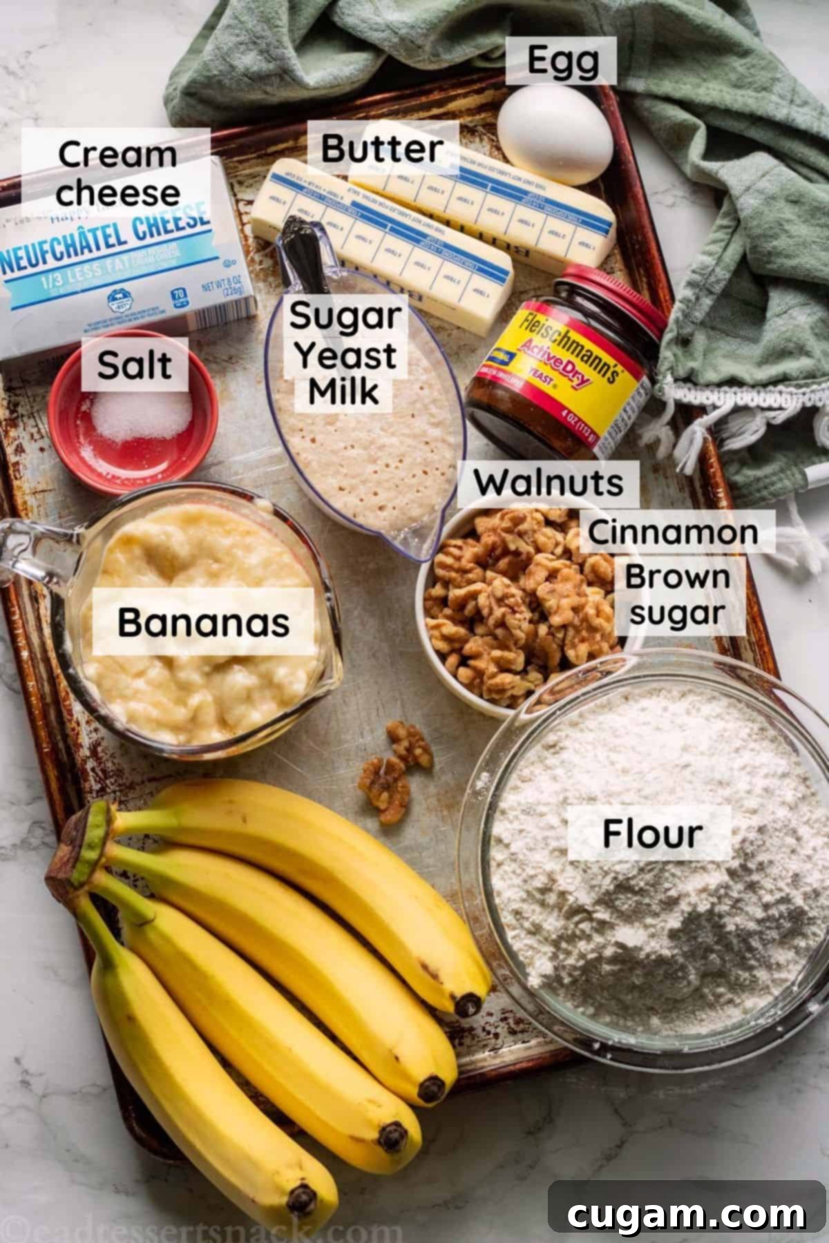 A baking sheet displays the fresh ingredients for making banana cinnamon rolls, including ripe bananas, butter, flour, and spices, alongside a green dish towel.