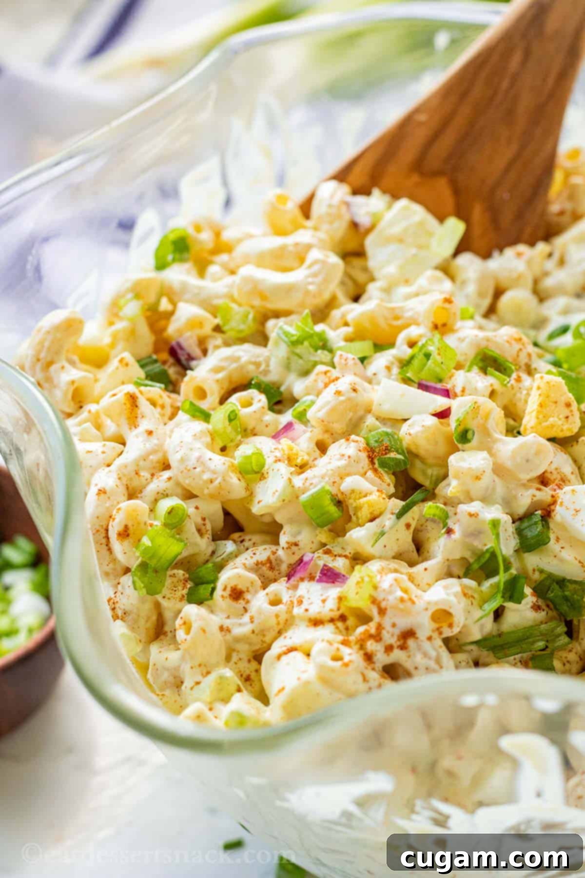 Chilled deviled egg macaroni salad in a beautiful glass dish with wavy edges, garnished with fresh green onions and a sprinkle of paprika, ready to be served with a wooden spoon.