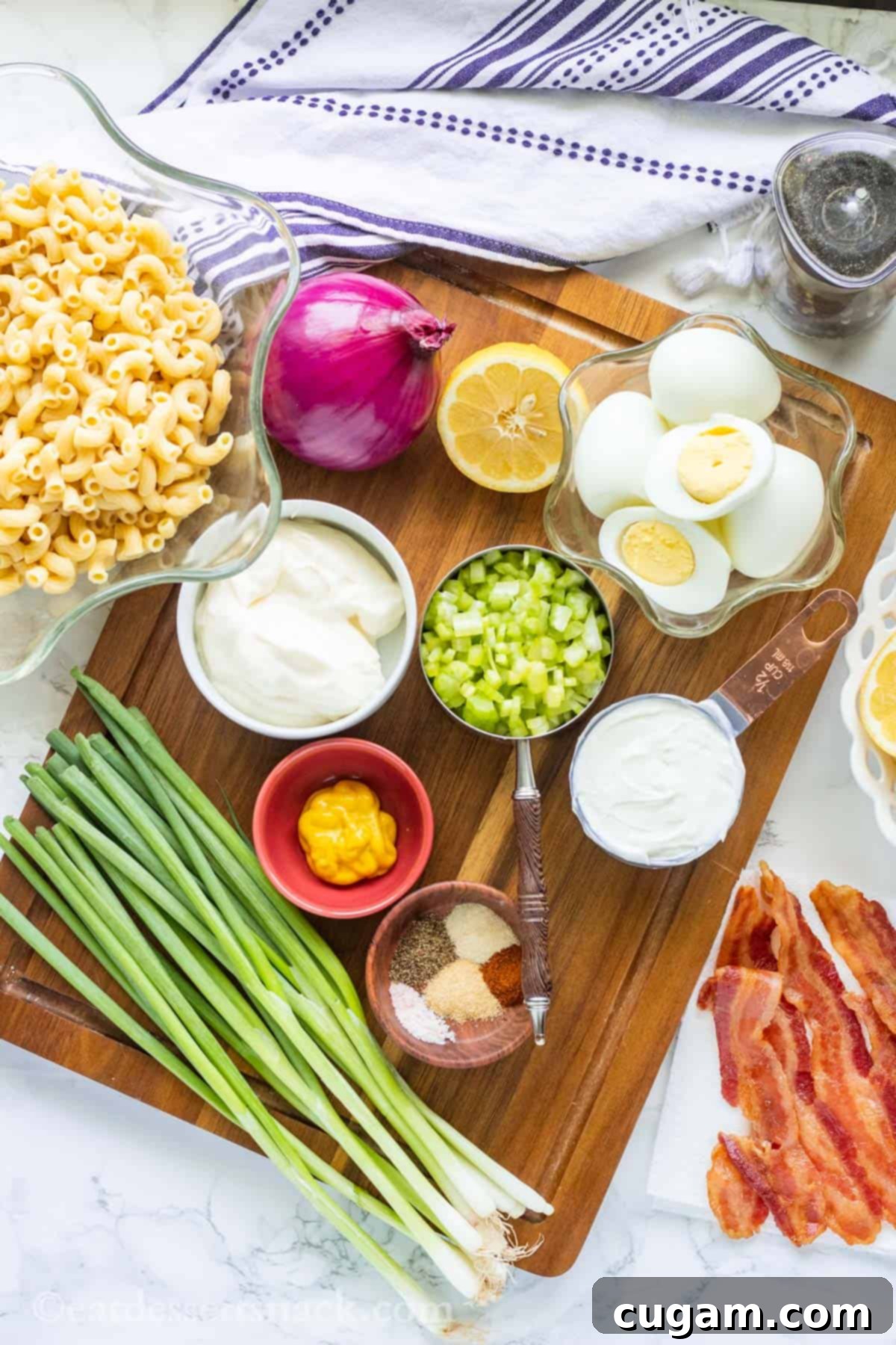 A beautiful arrangement of key ingredients for Deviled Egg Pasta Salad, including vibrant green onions, yellow mustard, various spices, freshly boiled eggs, creamy mayonnaise, sour cream, fresh lemon, red onion, and crispy bacon pieces, all artfully displayed on and around a rustic wood cutting board.