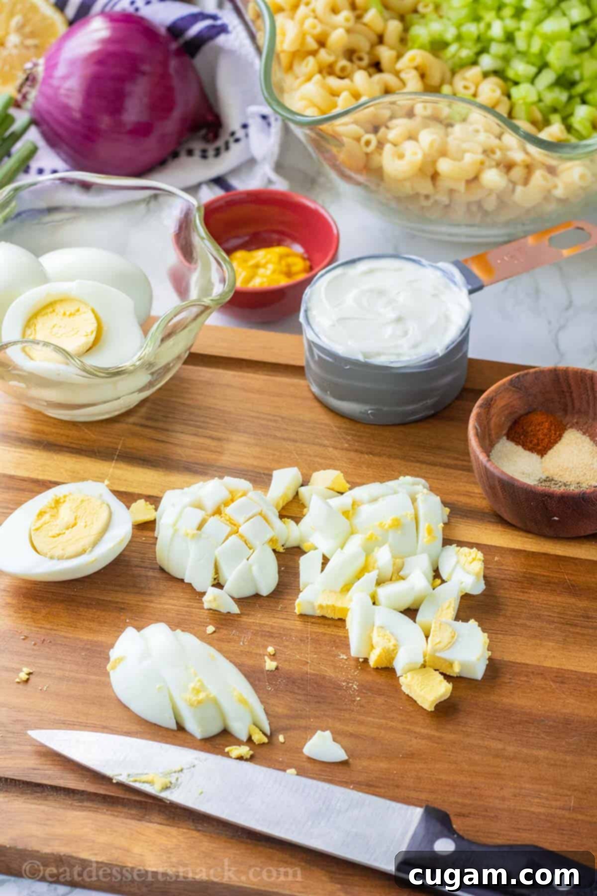 A close-up shot of hard-boiled eggs being expertly chopped on a rustic wooden cutting board, with various spices and a bowl of mayonnaise visible in the softly blurred background.
