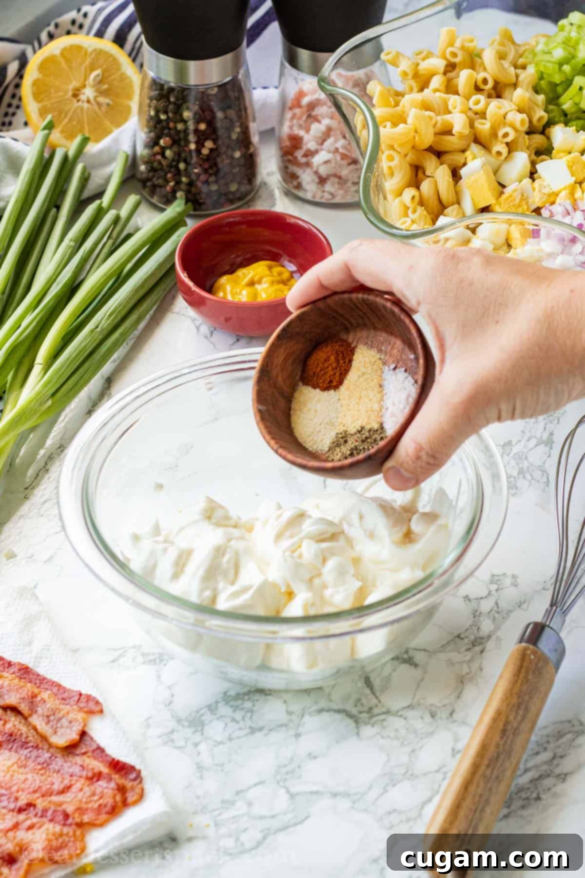 Hands carefully pouring a variety of aromatic spices into a clear glass bowl containing a creamy mixture of mayonnaise and sour cream, set on a elegant marble surface. In the background, fresh green onions, salt, pepper, and other chopped ingredients for the deviled egg salad are visible, creating a vibrant culinary scene.