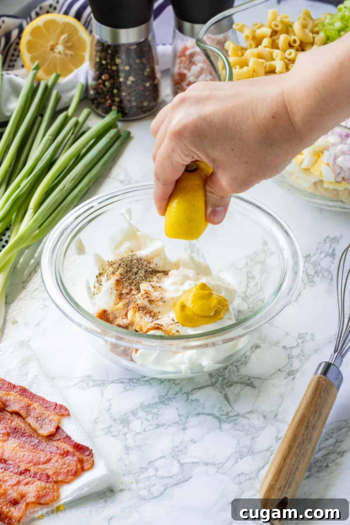 A hand is squeezing fresh lemon juice into a bowl filled with a creamy mixture of mayonnaise, mustard, sour cream, and various spices, all destined for the delicious deviled egg pasta salad.