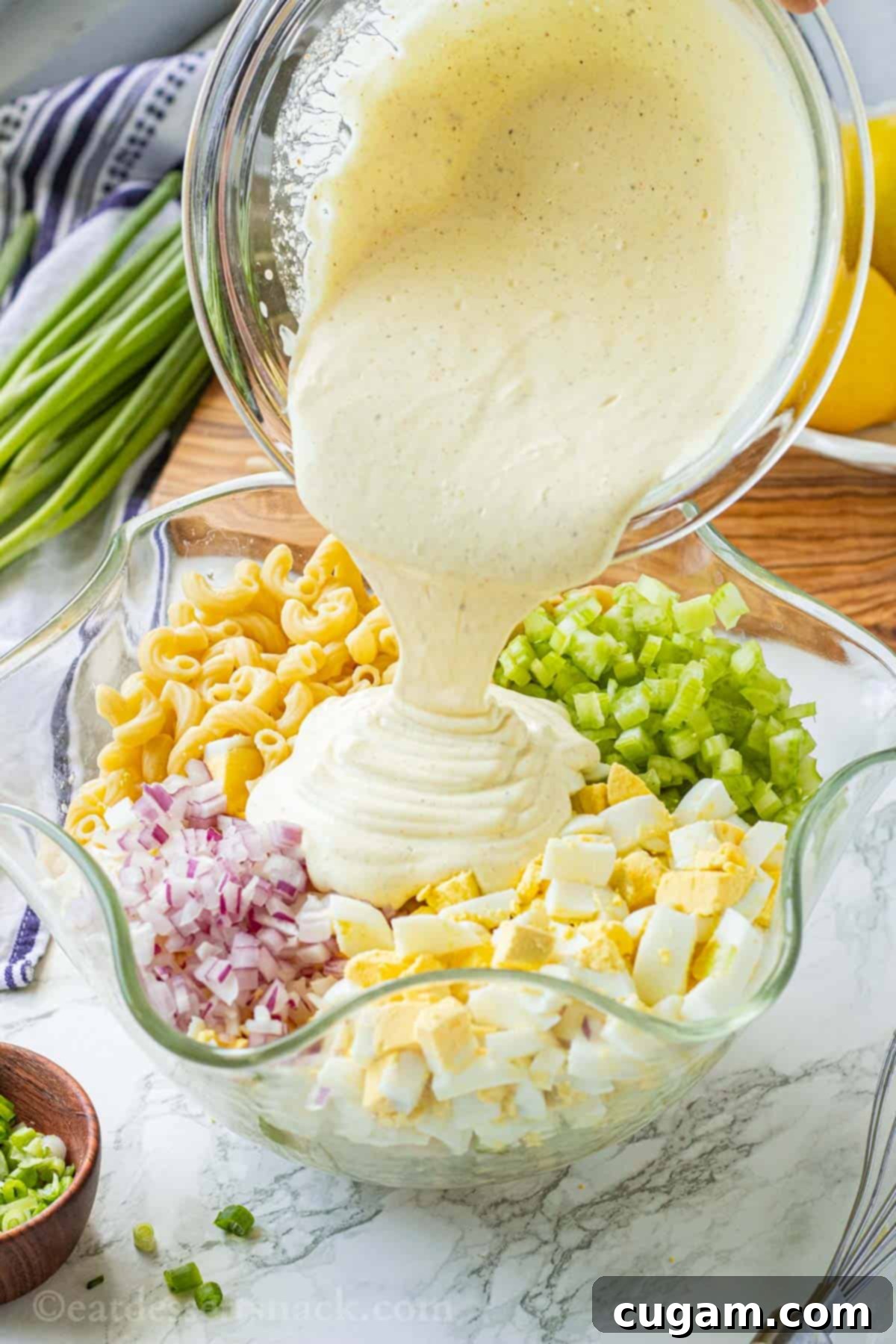 A delicious creamy dressing is being poured from a small bowl into a larger glass bowl with wavy edges, which is already filled with a vibrant mix of deviled egg pasta salad ingredients, ready for a gentle toss.