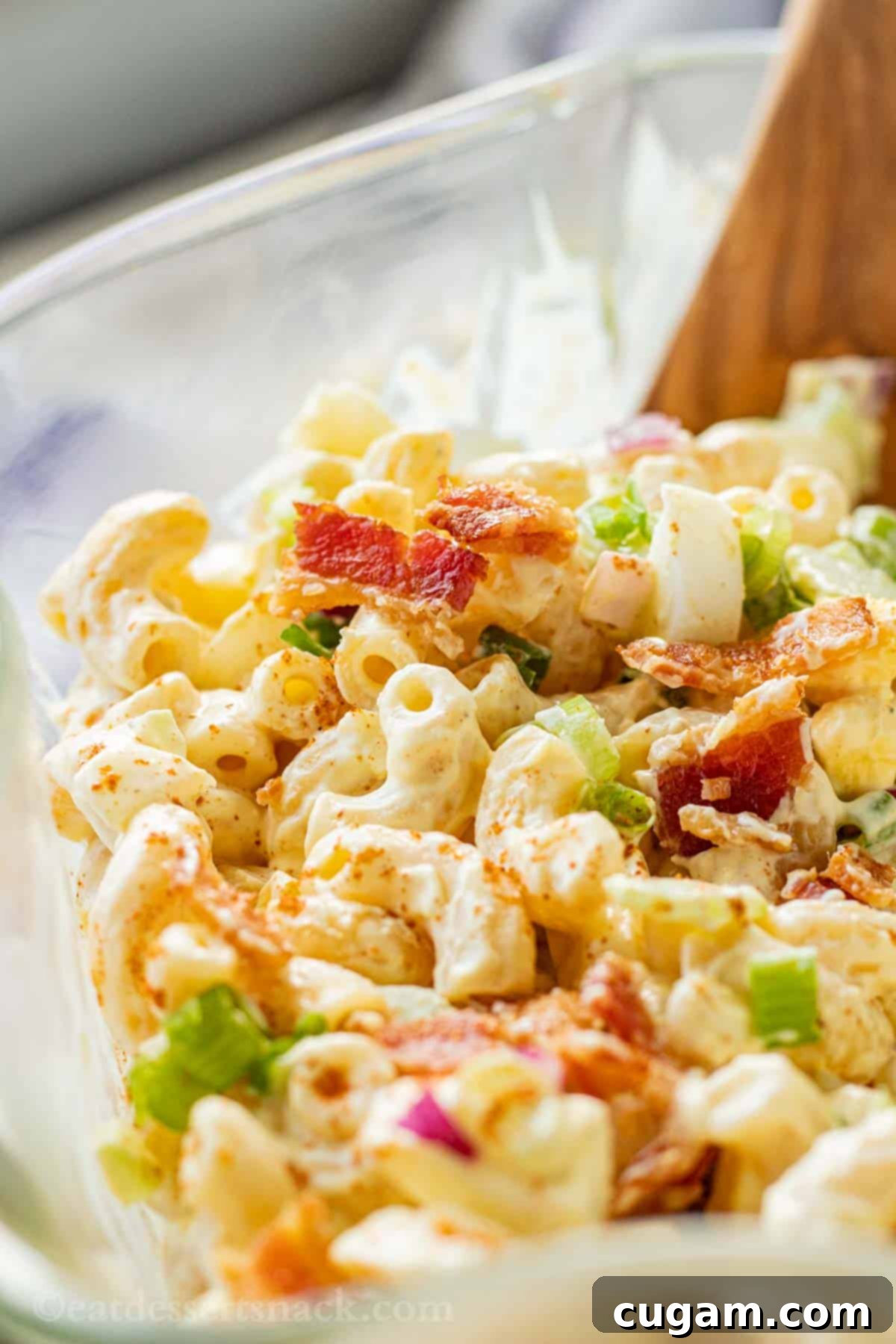 A generous serving of deviled egg macaroni salad, presented in a clear glass bowl with a rustic wooden spoon. The salad is beautifully garnished with crispy bacon pieces, highlighting its rich and inviting textures.