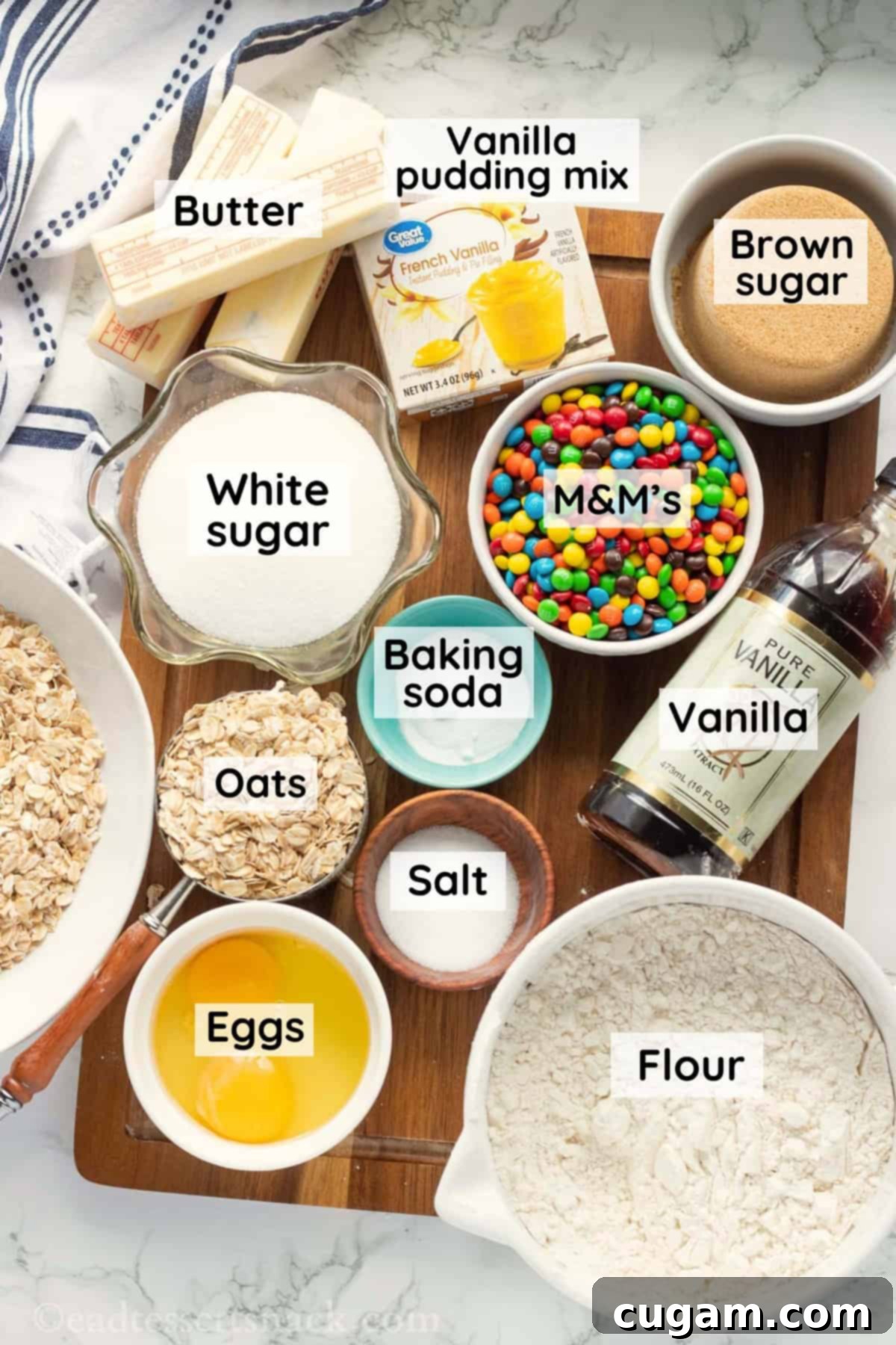 Labeled ingredients for oatmeal m&m cookies on wood cutting board on marble surface.