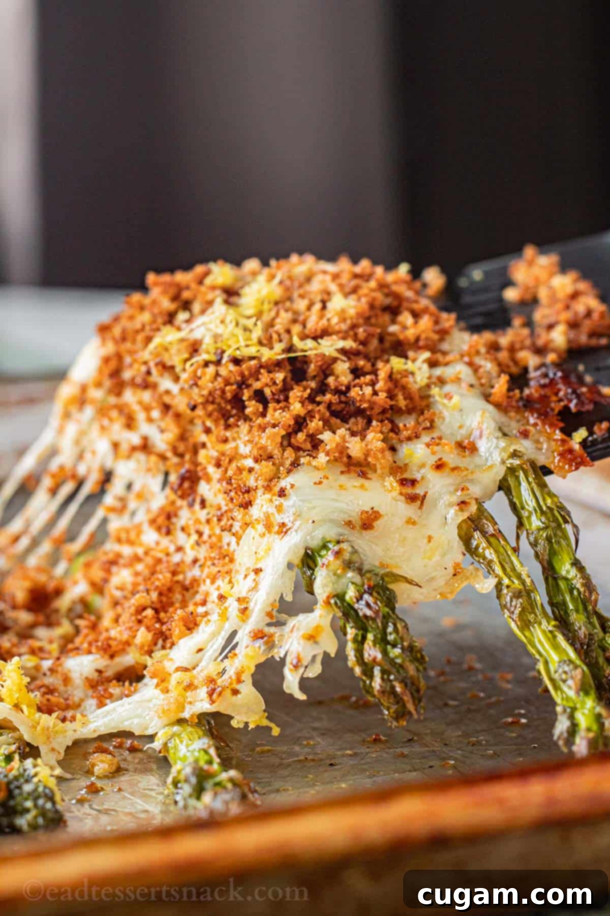 A spatula lifting a piece of baked cheesy asparagus with melted cheese stretching, off a metal baking sheet.