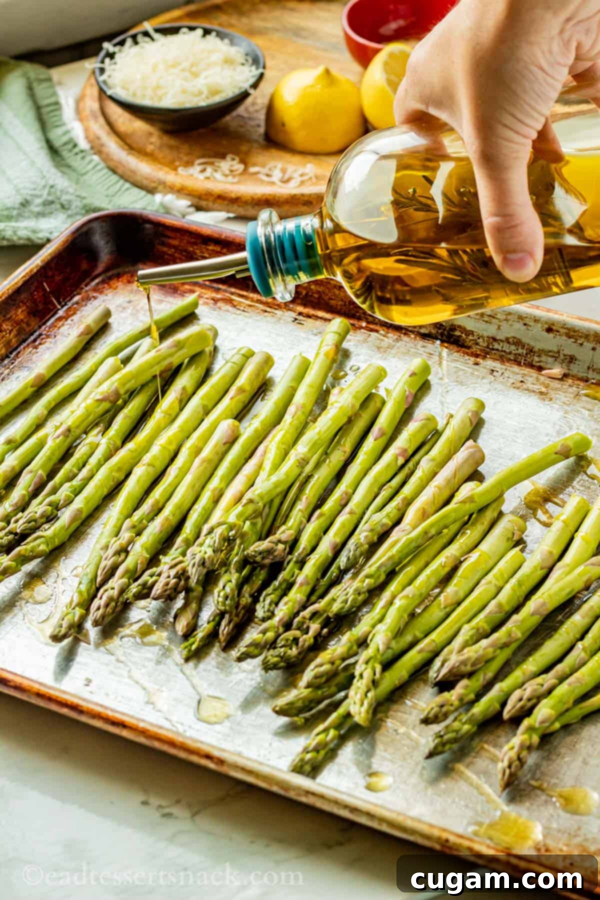 Hand drizzling olive oil from a bottle over raw asparagus spears on a metal baking pan.