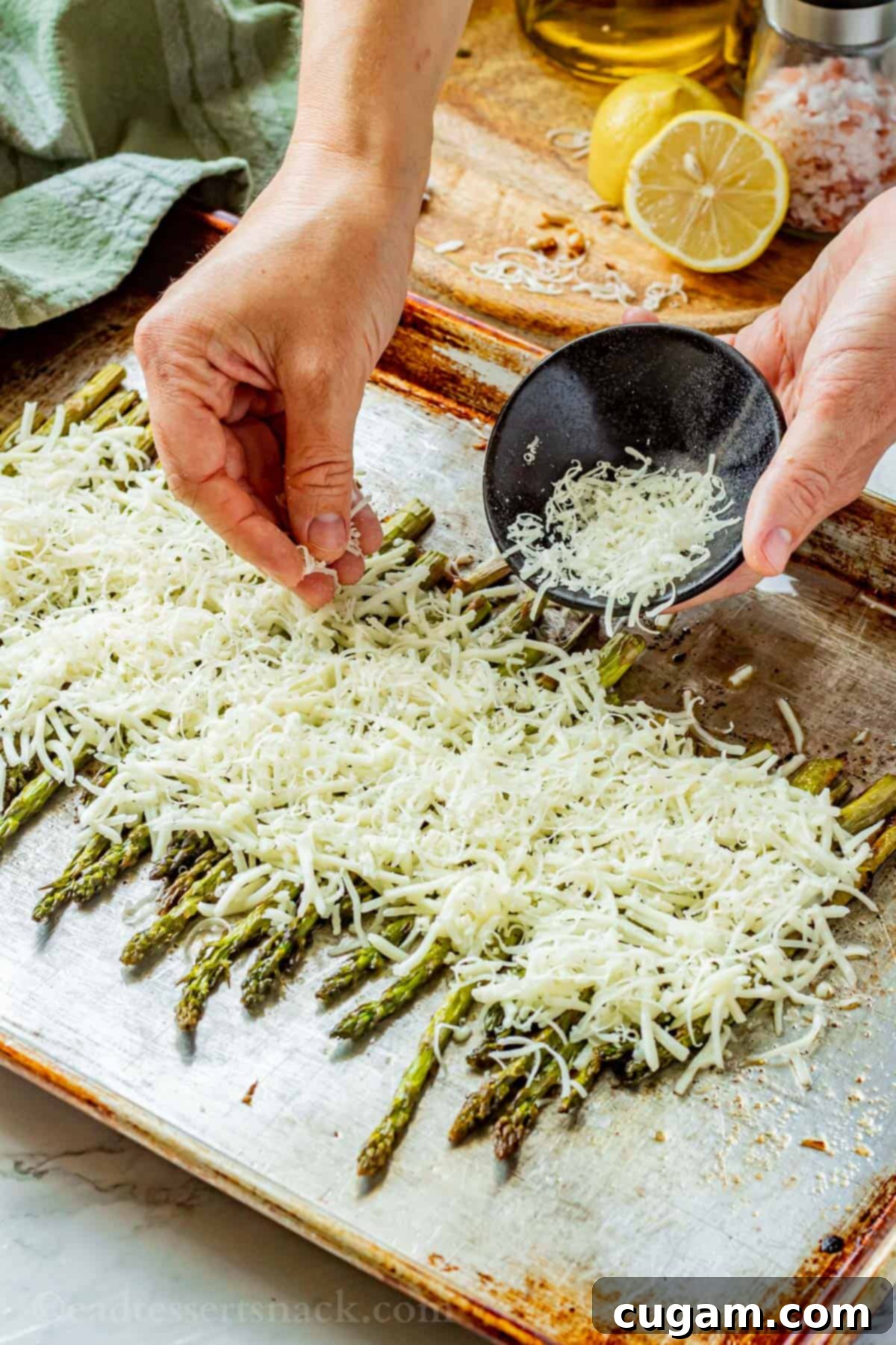 Hand sprinkling grated cheese over baked asparagus on a metal baking pan, fresh out of the oven.