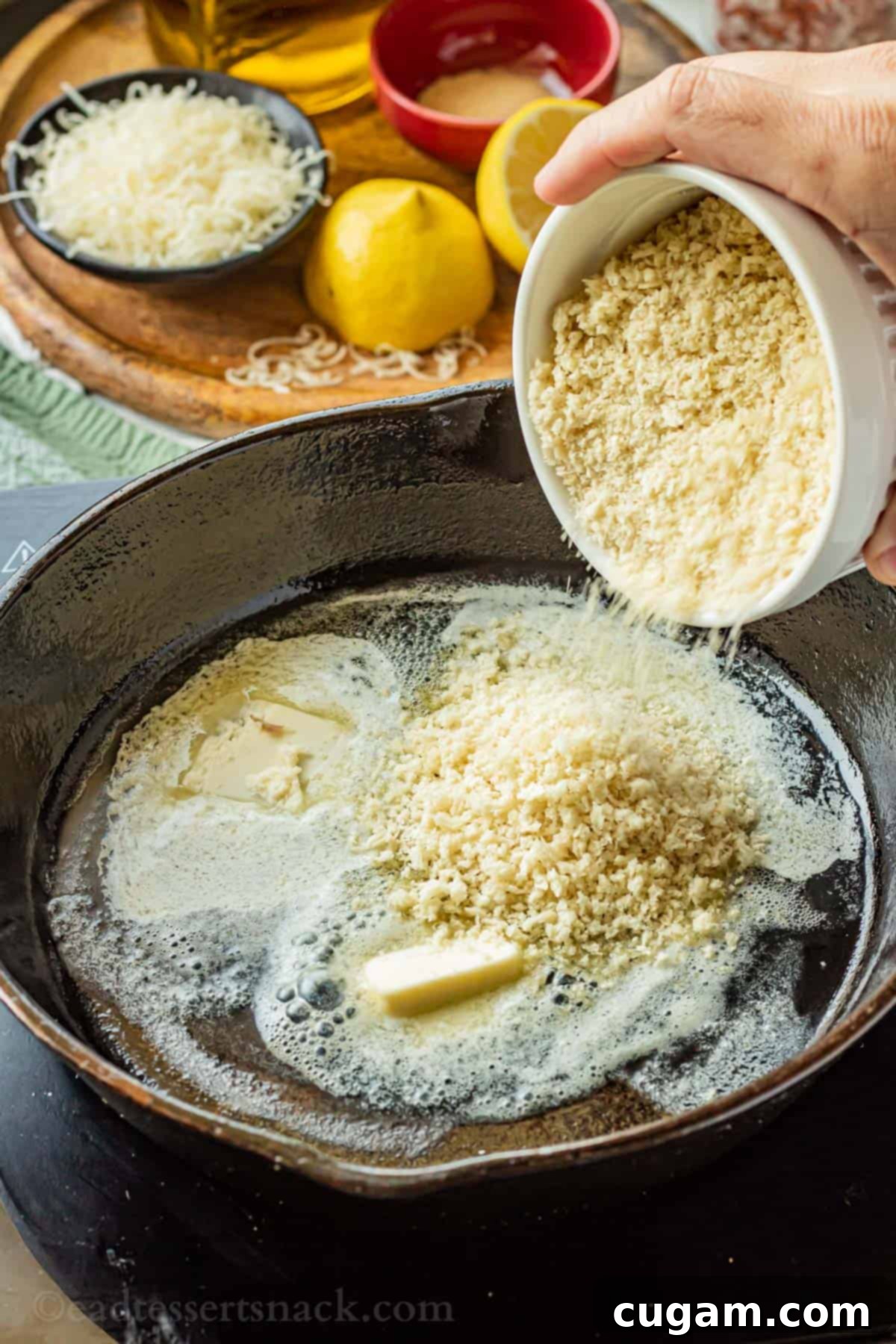 Pouring panko breadcrumbs into a black frying pan containing melting butter on a stovetop.