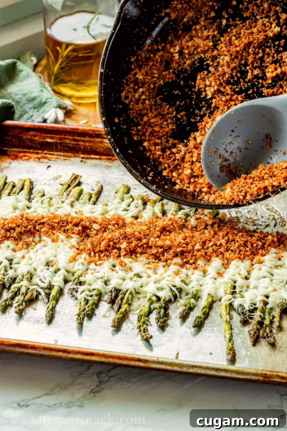 A spoon spreading golden toasted panko breadcrumbs over a metal pan of baked cheesy asparagus.