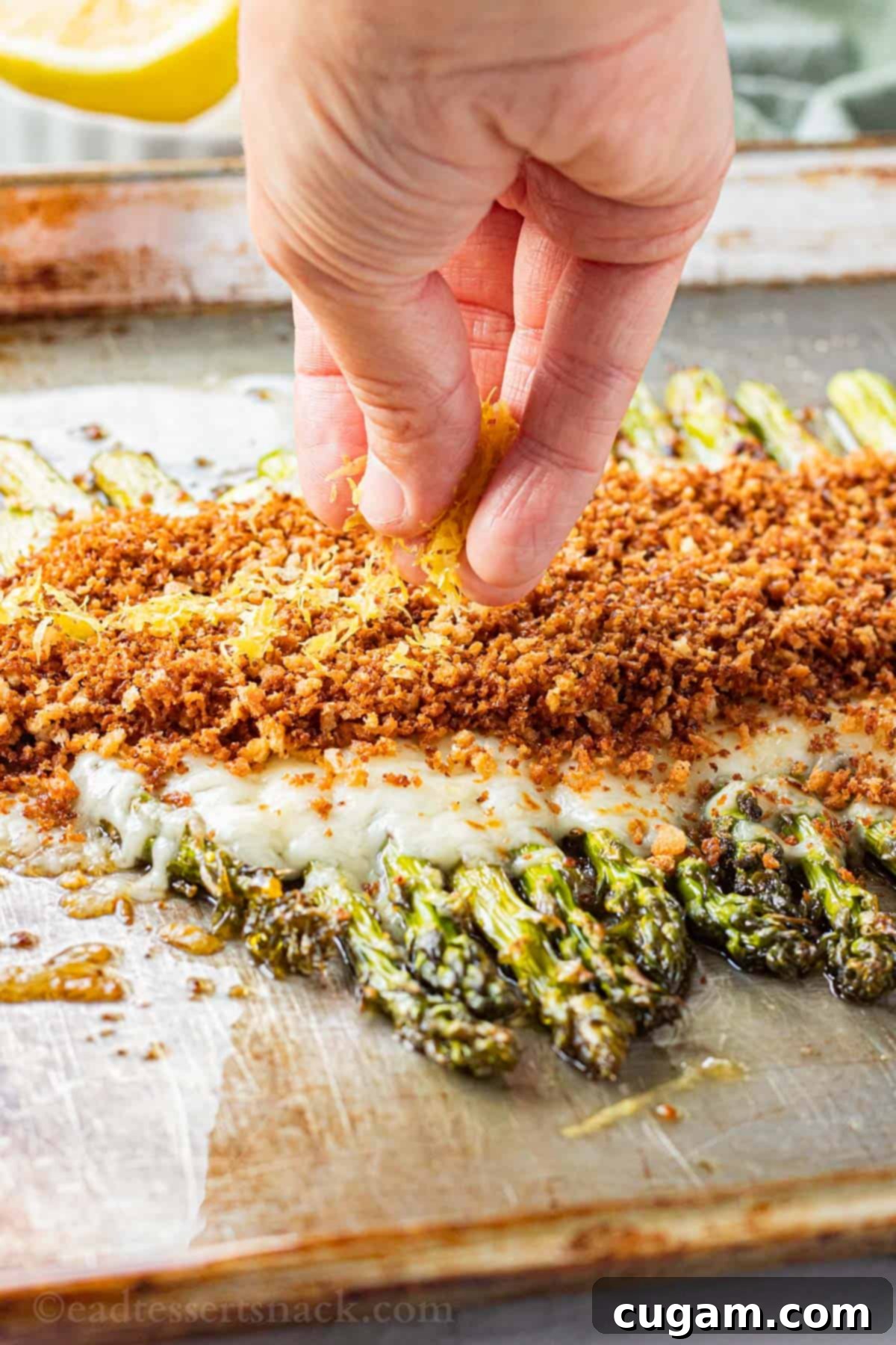 A hand sprinkling fresh lemon zest over baked cheesy asparagus topped with panko breadcrumbs.