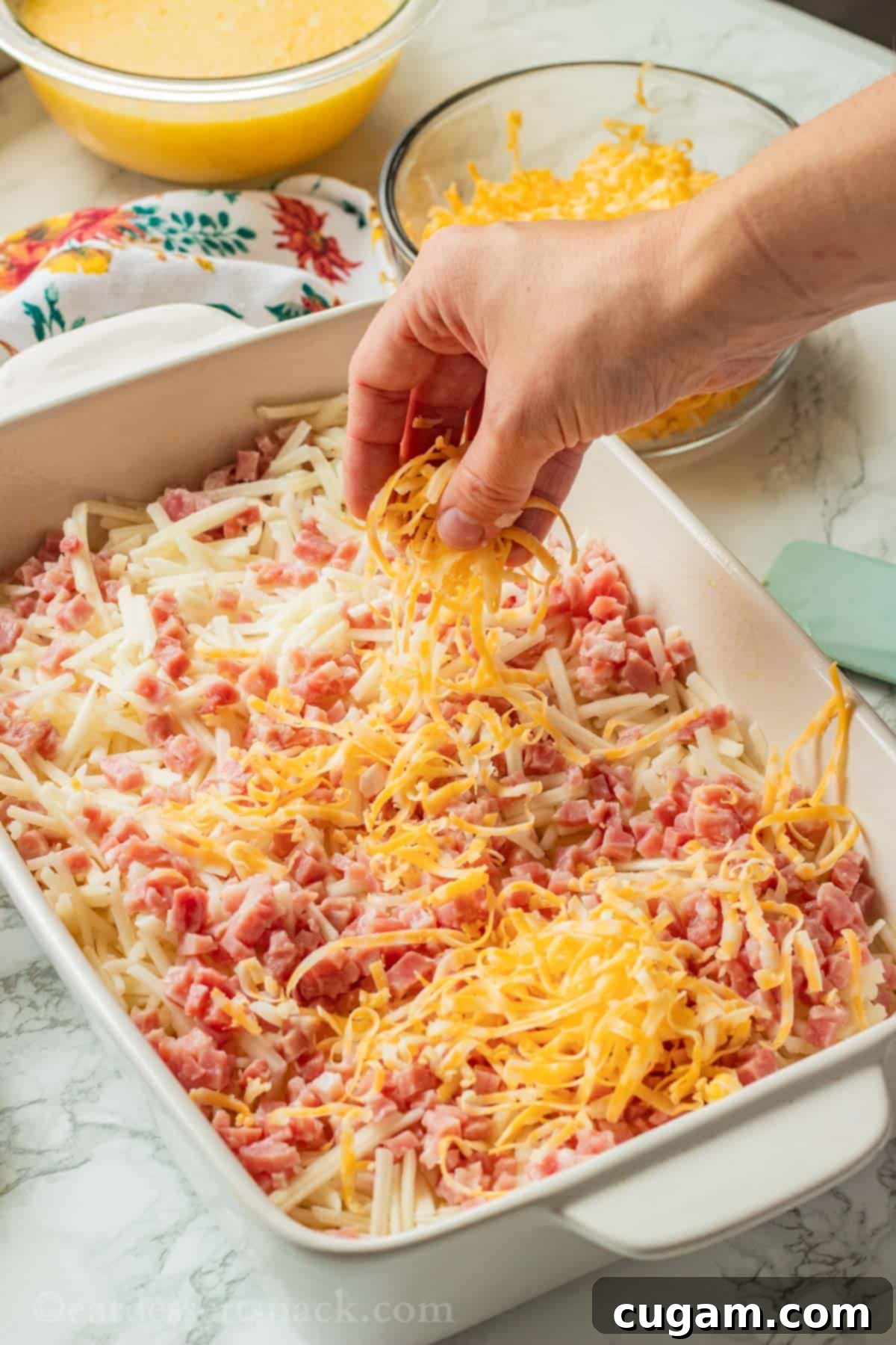 Hand sprinkling cheese over raw ham and cheese hash brown casserole