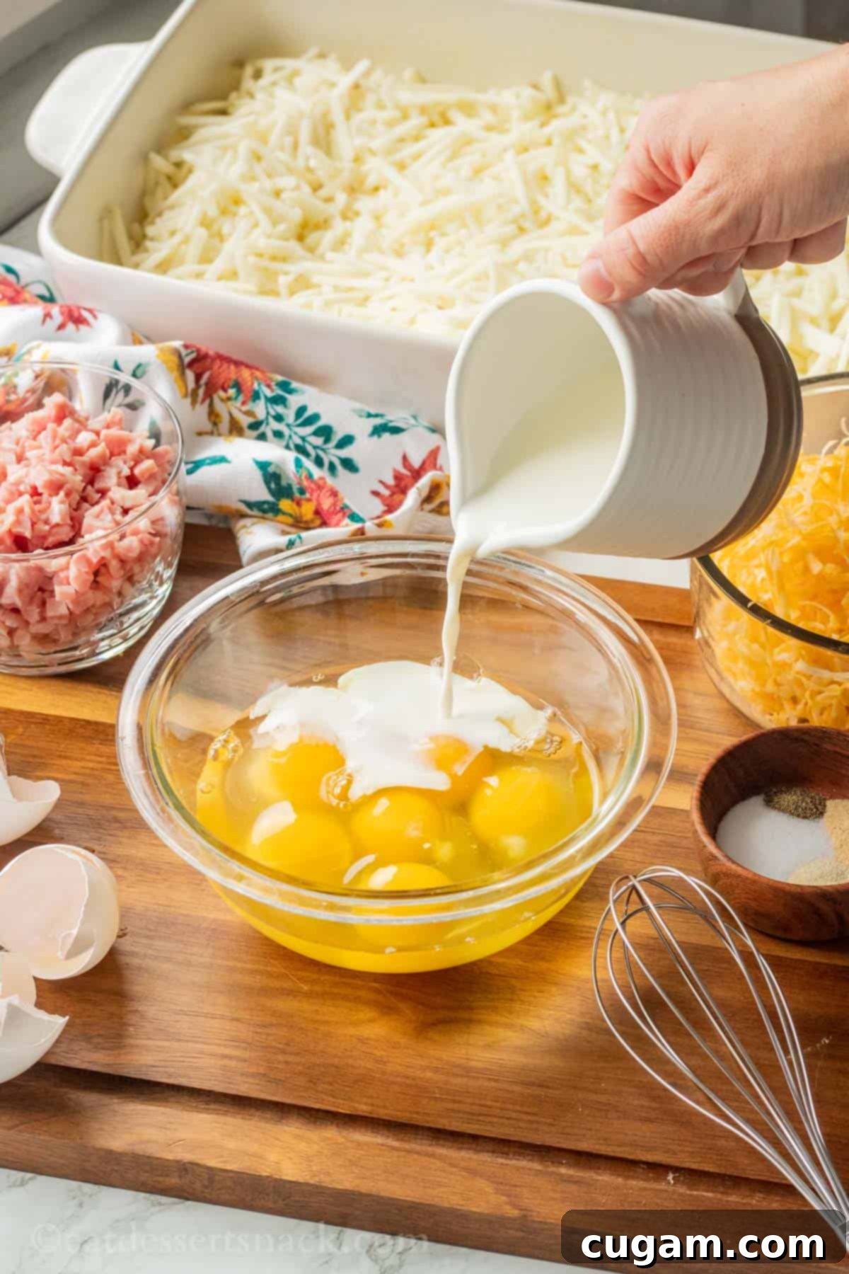 Hand pouring milk into glass bowl of eggs sitting on wood cutting board.