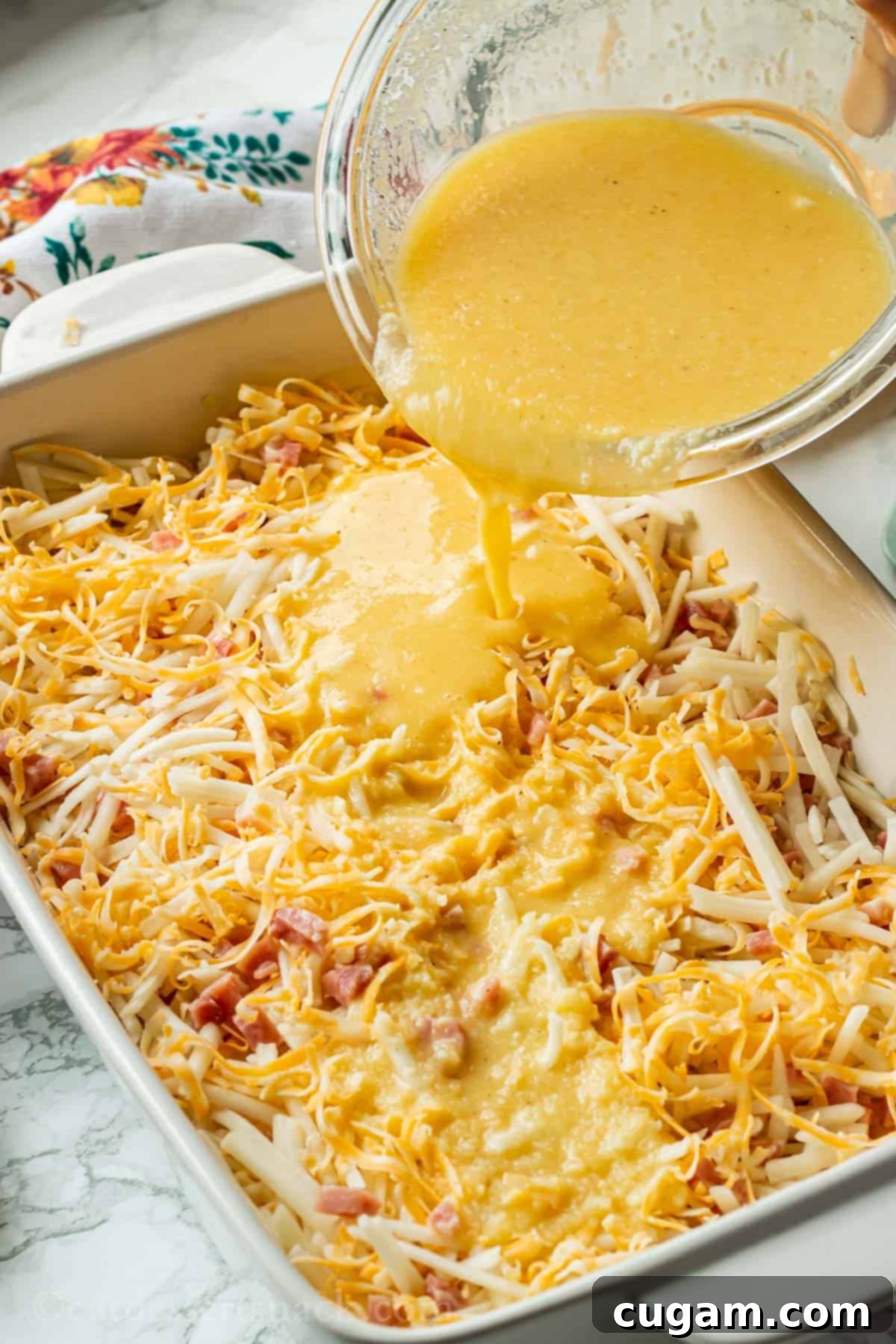 Hand pouring egg and butter mixture over white casserole dish of cheese, ham and hash brown potatoes.