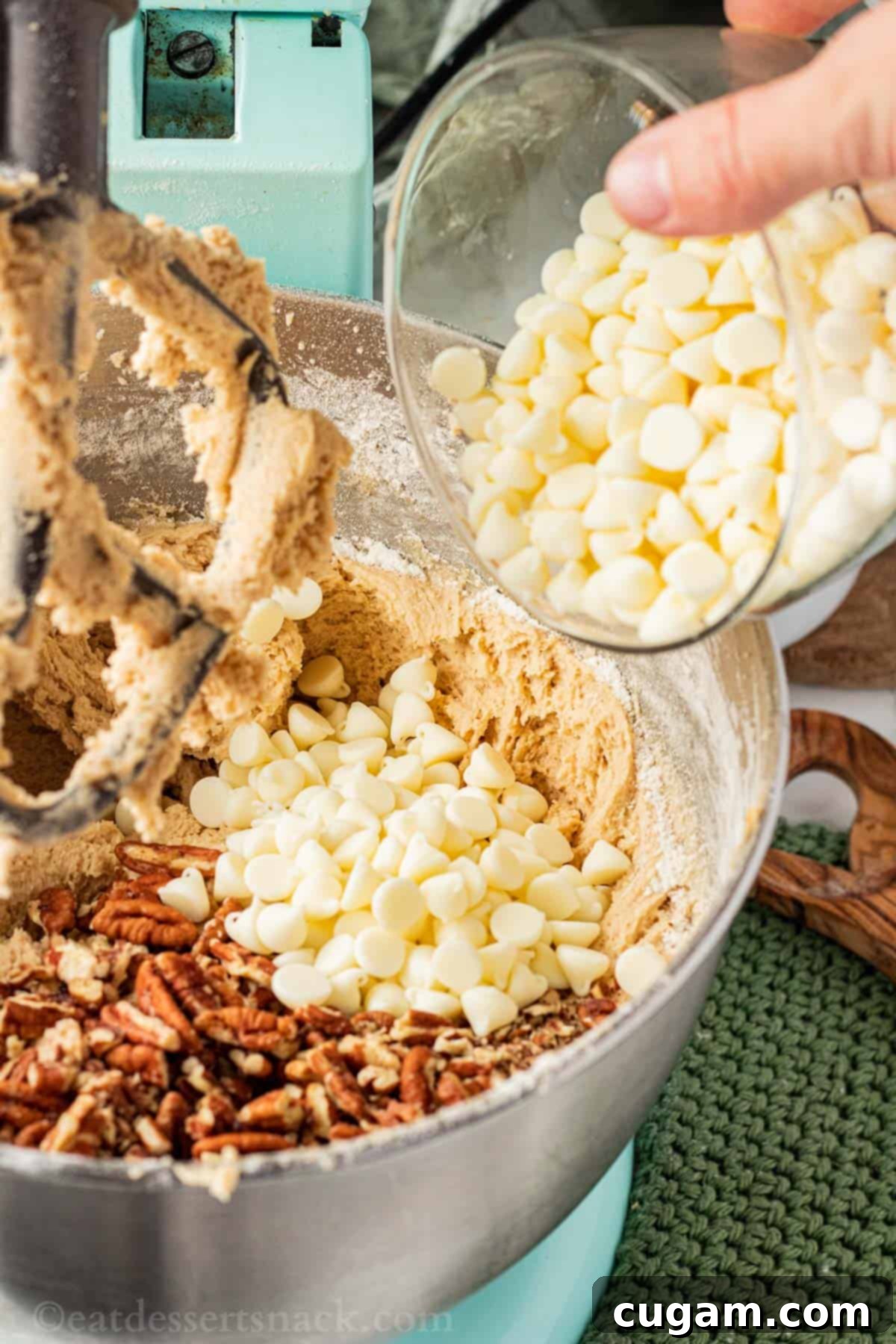 Hand pouring white chocolate chips into mixing bowl of raw maple cookie dough.