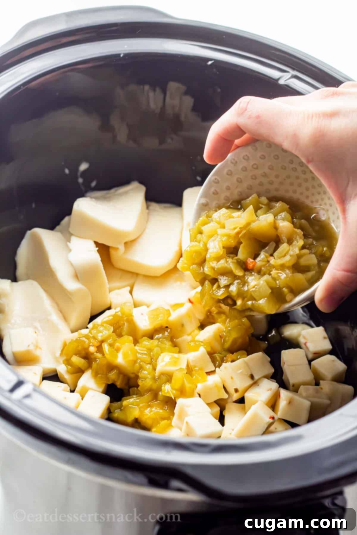 A hand pouring diced green chiles from a can into a black slow cooker, where blocks of cream cheese and white Velveeta cheese are already waiting, ready for the queso to melt.