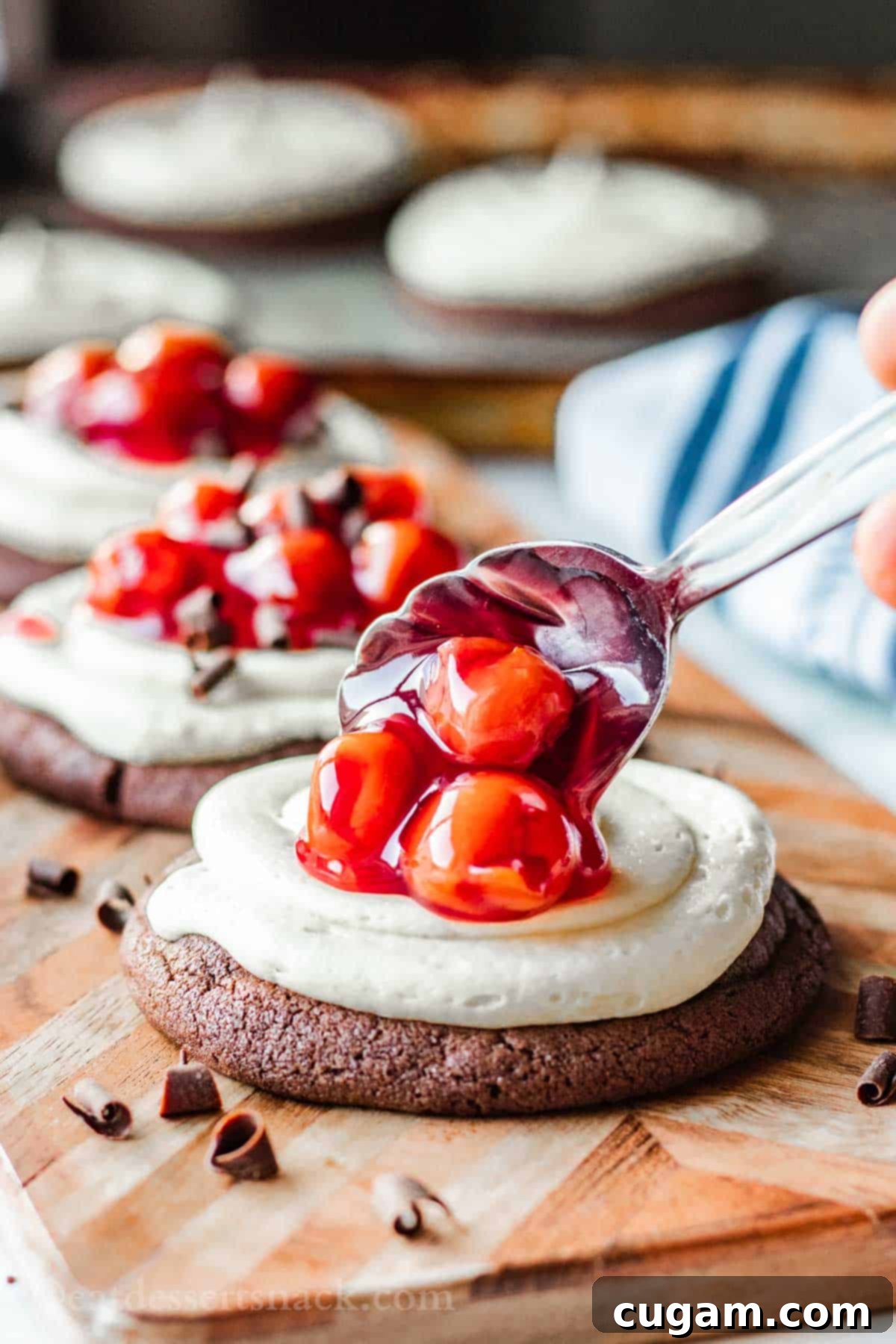 Spooning cherry pie filling on top of baked chocolate cookie with cheesecake frosting sitting on wood cutting board.