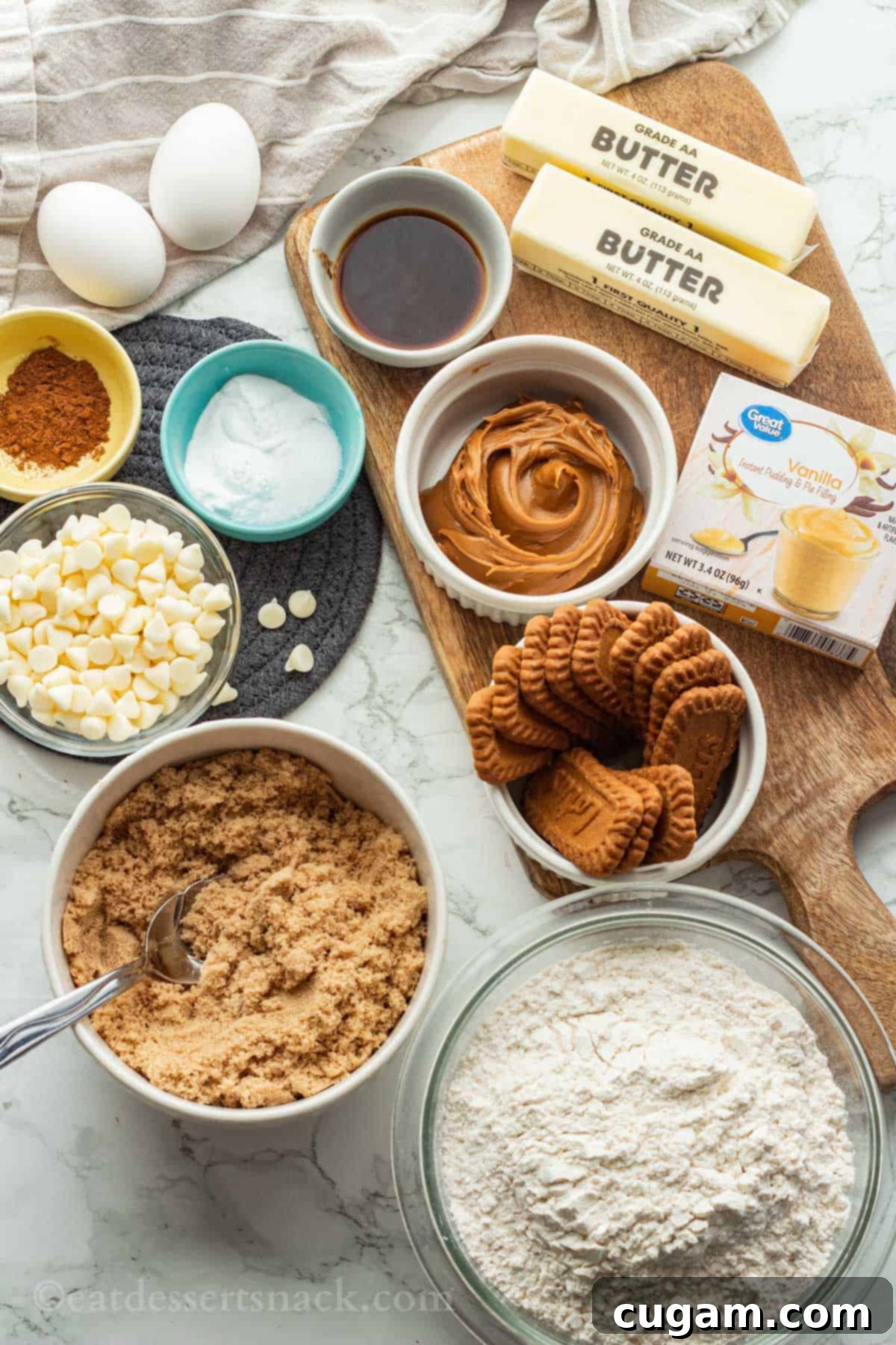 Ingredients for white chocolate biscoff cookies laid out on a marble and wood cutting board, including butter, eggs, cookie butter, and chocolate chips.