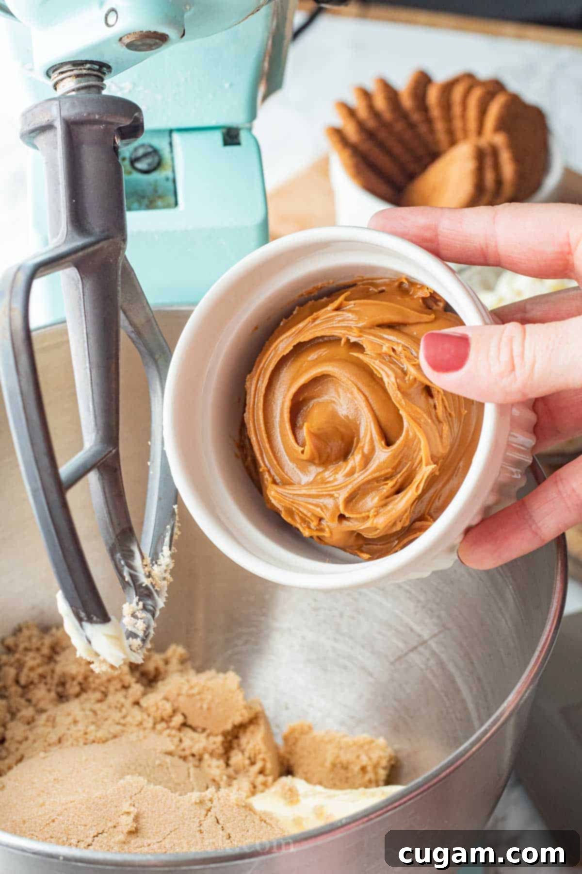Smooth cookie butter is scooped from a white bowl and poured into a metal mixing bowl with other ingredients for the cookie dough.