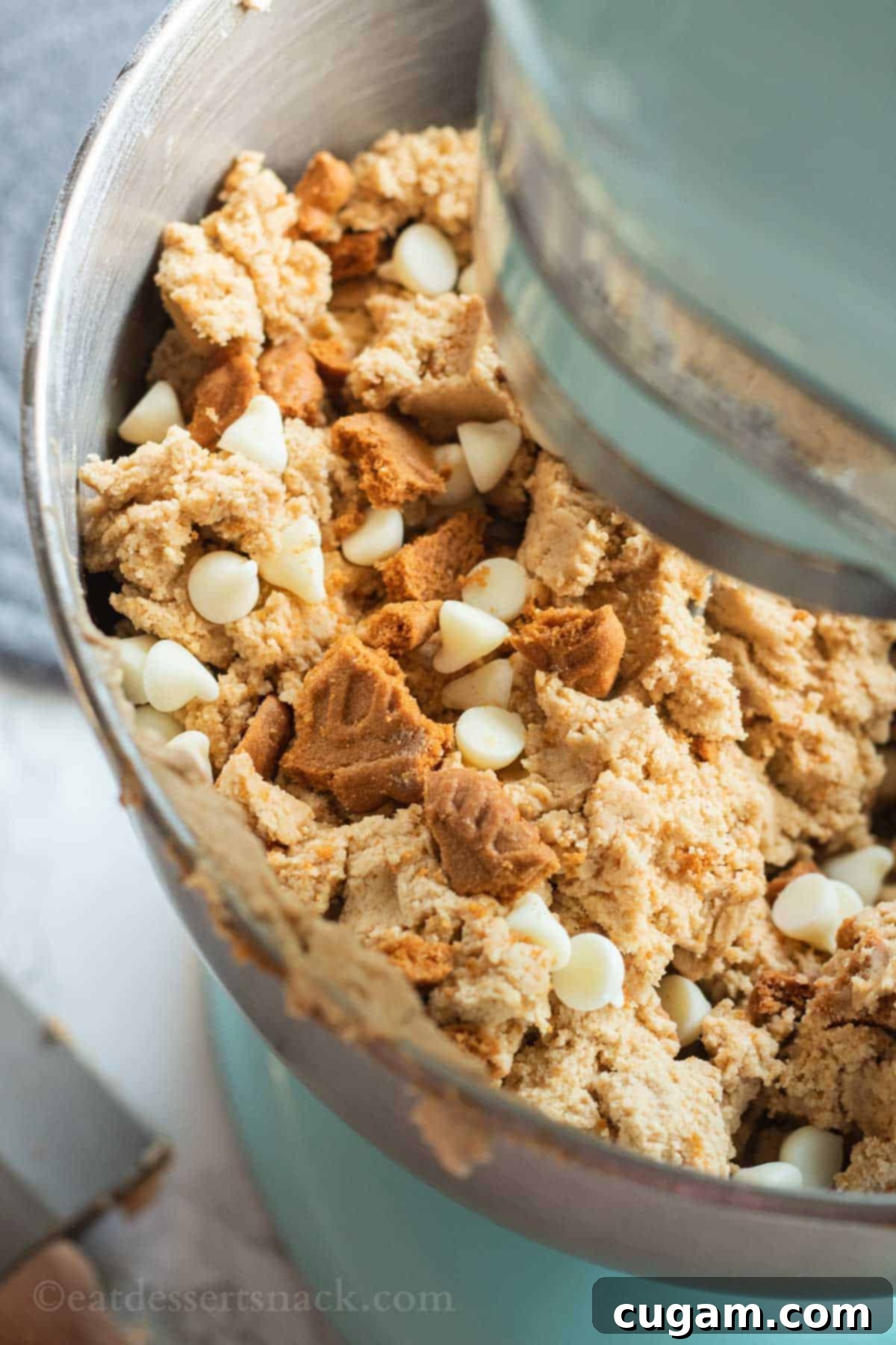 Raw white chocolate biscoff cookie dough sits in a metal bowl attached to a blue stand mixer, ready for baking.